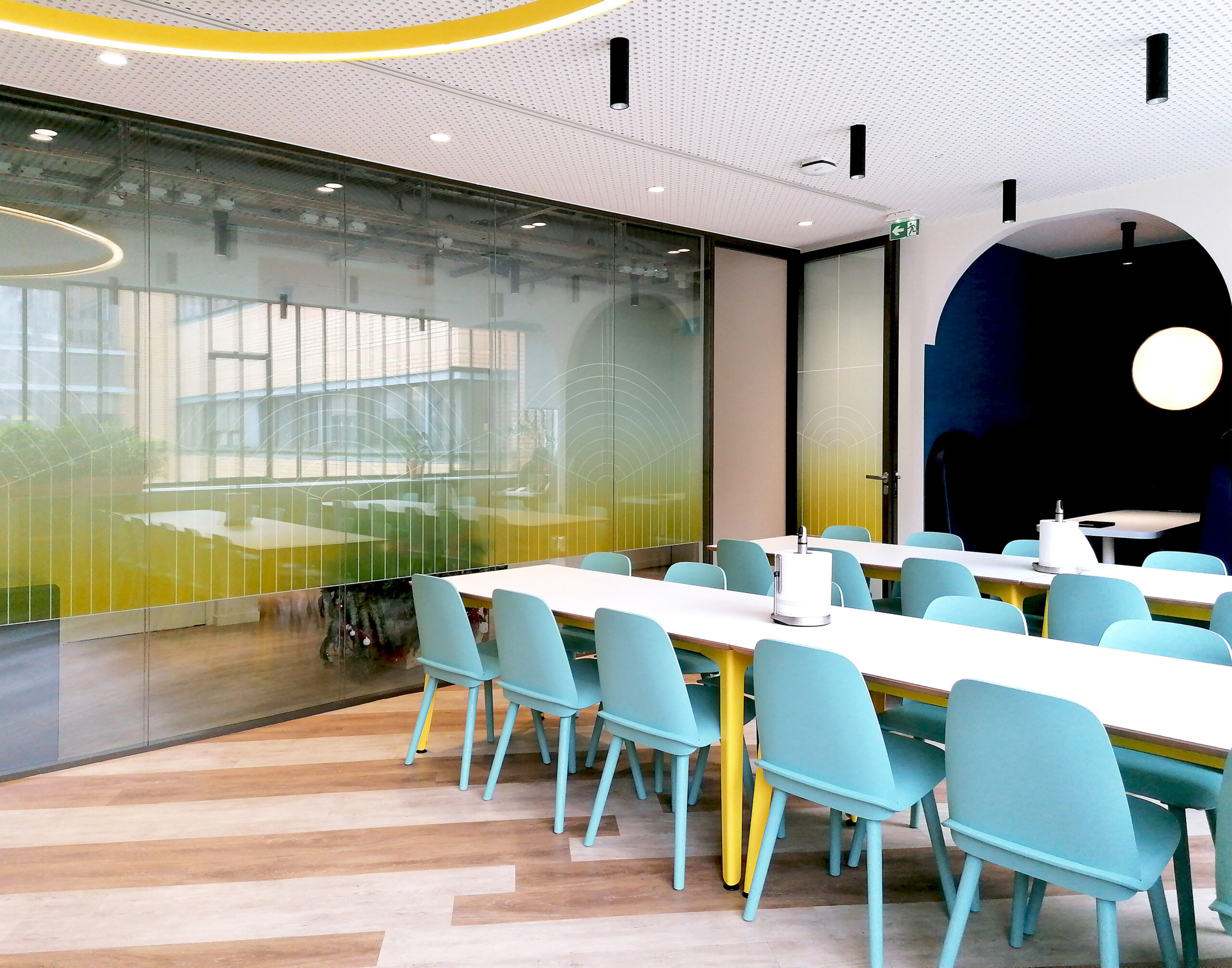 A modern conference room featuring a long white table surrounded by teal and yellow chairs. Glass walls allow natural light to filter in, complemented by abstract wall graphics. The design emphasizes collaboration and creativity, with a minimalist aesthetic and vibrant color palette enhancing the inviting atmosphere.