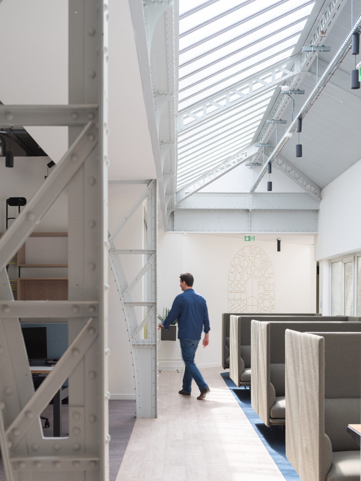 Light floods the spacious open-plan office through a glass skylight, highlighting the exposed metal beams and contemporary design. Cozy workstations with high-backed booths provide privacy, while a sleek, minimalist aesthetic is maintained throughout. A modern, neutral color palette enhances the airy atmosphere, promoting creativity and collaboration.