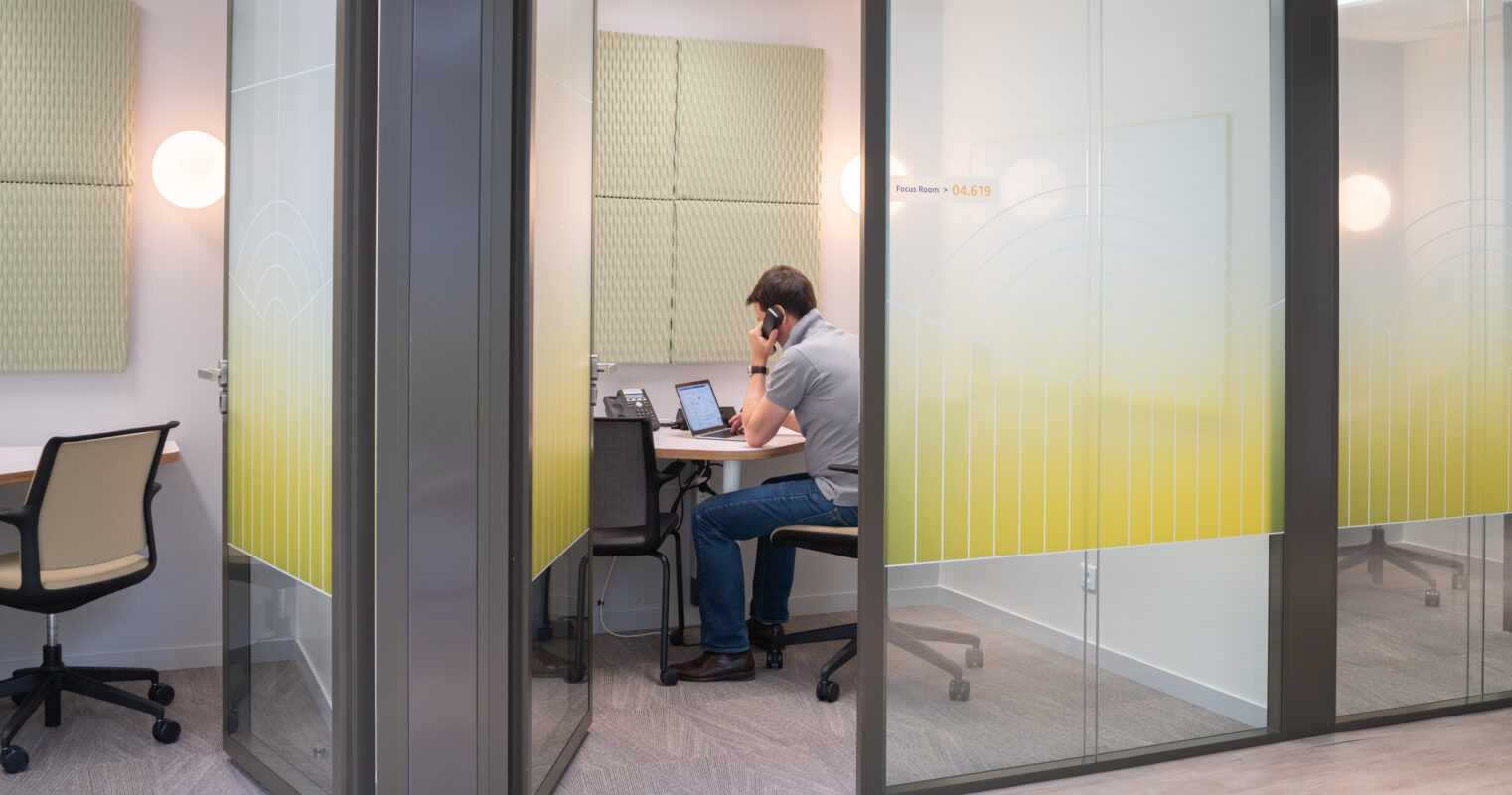 A modern office workspace features a man seated at a sleek, minimalist desk with a laptop and headset. Sound-absorbing wall panels add acoustic comfort, while large glass partitions enhance natural light. Subtle yellow gradient graphics on the glass maintain privacy while fostering an open, collaborative atmosphere.