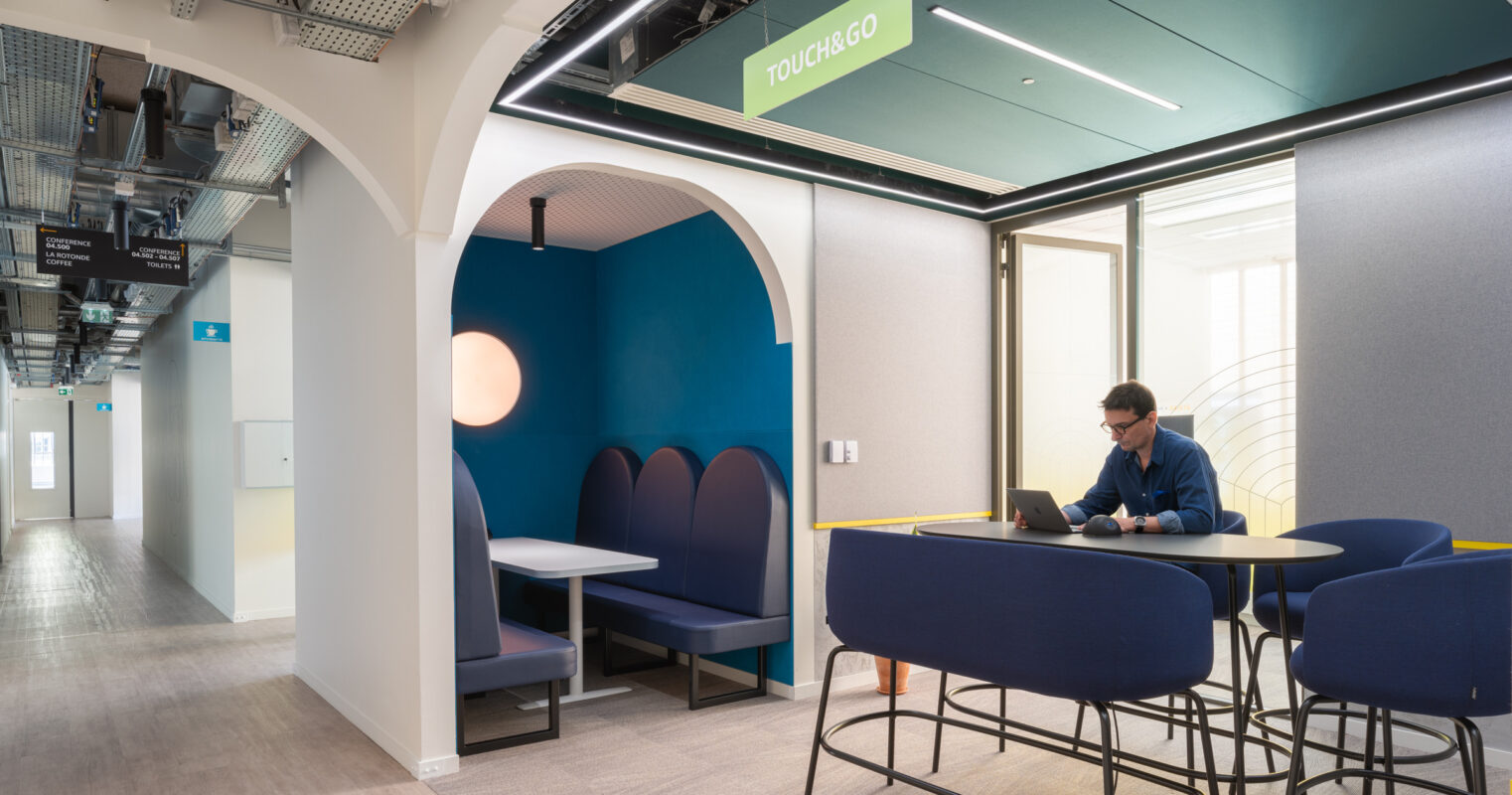 A modern collaborative workspace featuring a cozy blue booth and round table. Natural light filters through a large window, illuminating the contemporary design elements, including arched architectural details and vibrant colors. A professional sits at the table, engaged with a laptop, emphasizing functionality and comfort in the environment.