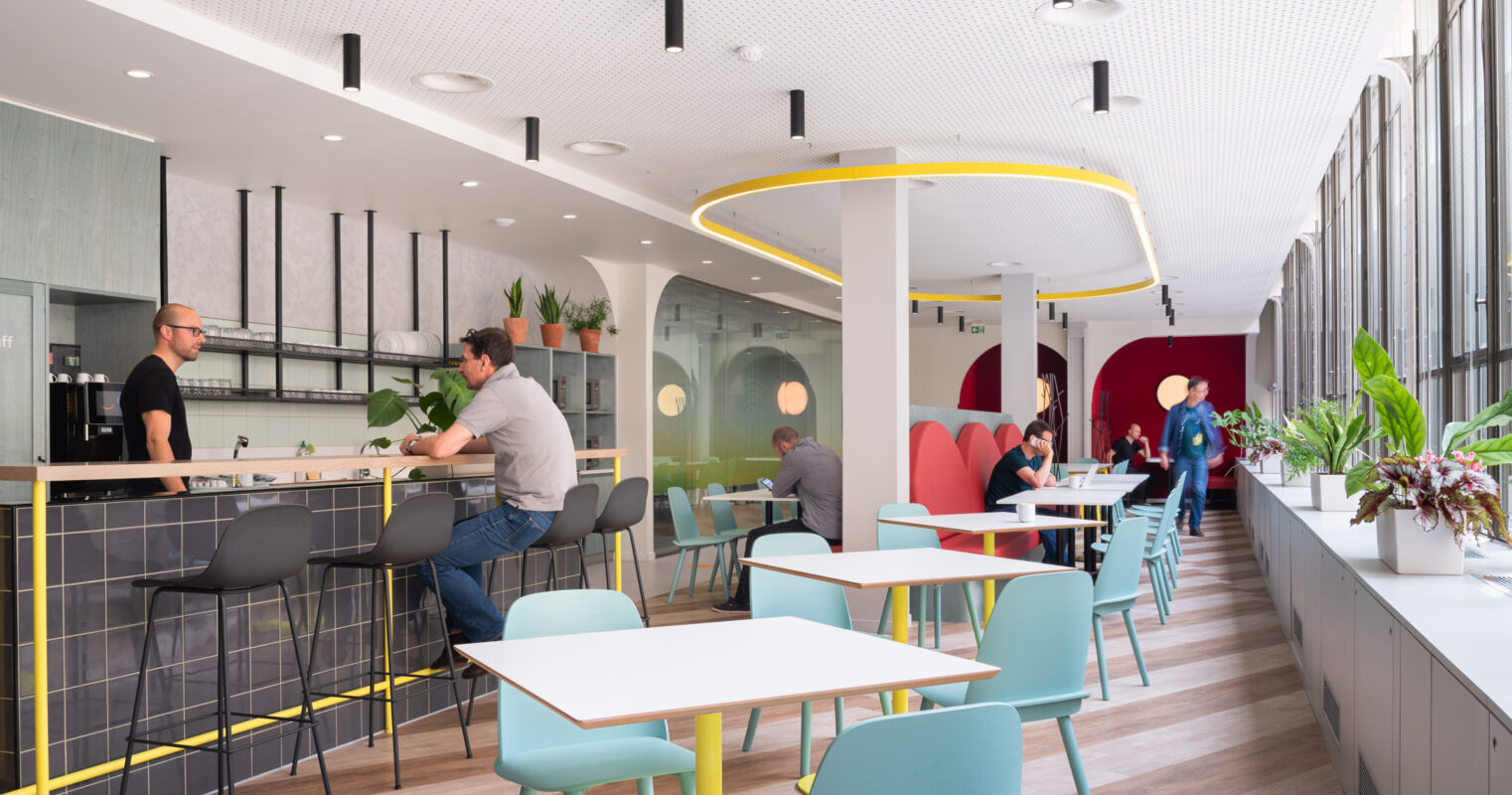 A modern café interior features sleek, minimalist furniture with pastel-colored chairs and light wooden flooring. A bright bar area with a marble counter offers seating along one side, while patrons enjoy the lively atmosphere surrounded by greenery. Circular light fixtures and bold wall colors create a vibrant ambiance.