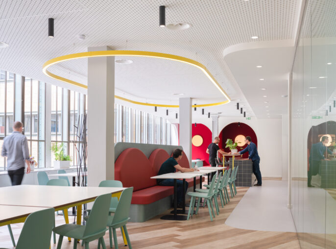 Modern office interior showcasing a vibrant, collaborative workspace. The design features a mix of comfortable booth seating in red tones, light wooden flooring, and large windows allowing natural light. Unique circular ceiling lights and colorful accents create an inviting atmosphere, promoting creativity and interaction among employees.