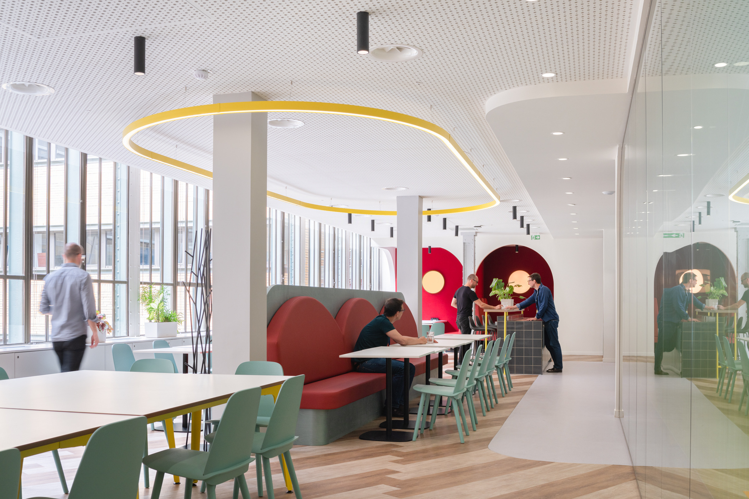 Modern office interior showcasing a vibrant, collaborative workspace. The design features a mix of comfortable booth seating in red tones, light wooden flooring, and large windows allowing natural light. Unique circular ceiling lights and colorful accents create an inviting atmosphere, promoting creativity and interaction among employees.