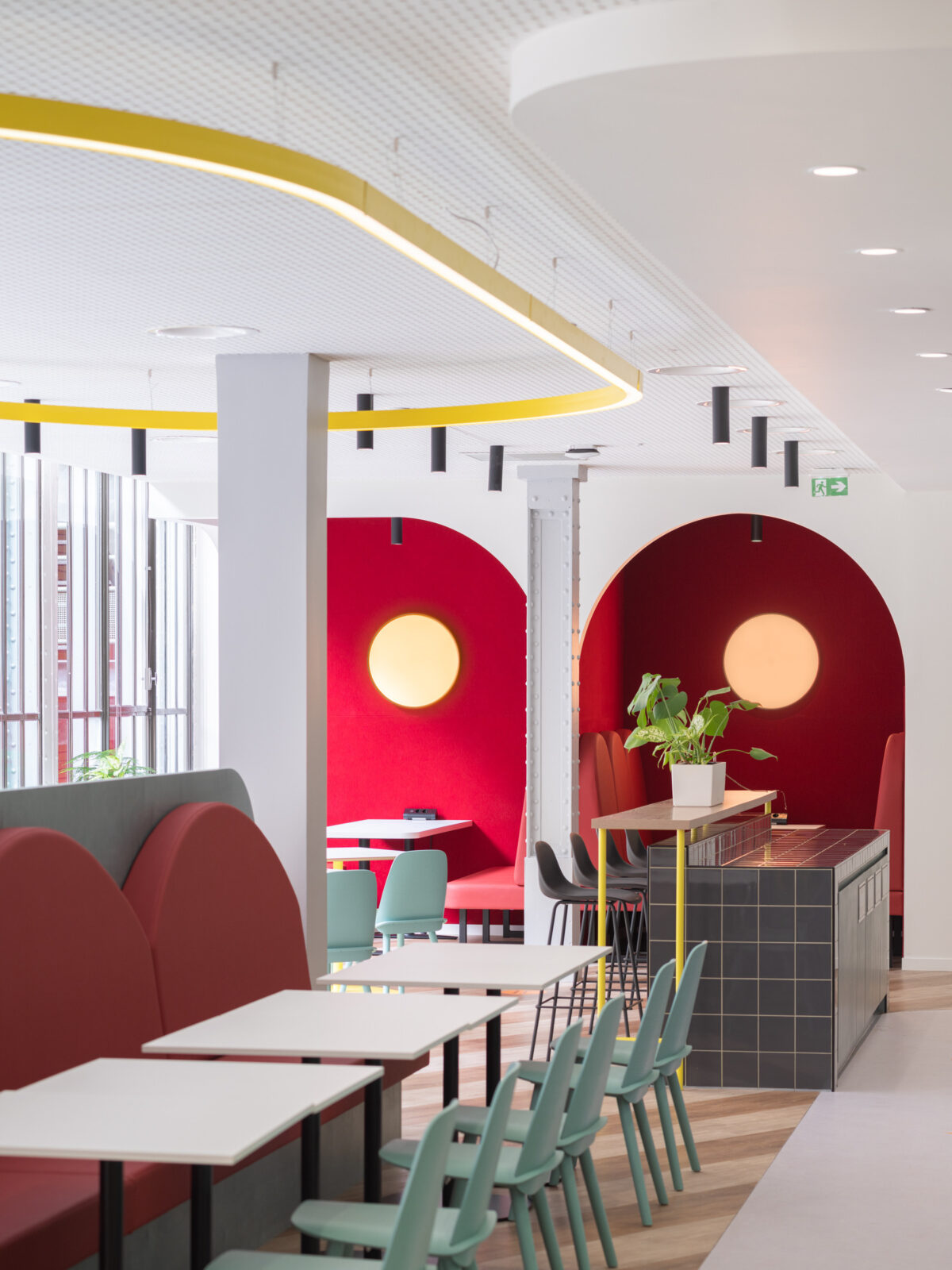 A modern café interior showcasing curved architectural features with vibrant red accent walls and circular light fixtures. Stylish, soft seating in muted tones complements the sleek, minimalist furniture. The open layout encourages social interaction, while carefully placed plants enhance the inviting atmosphere, creating a lively yet cozy space.