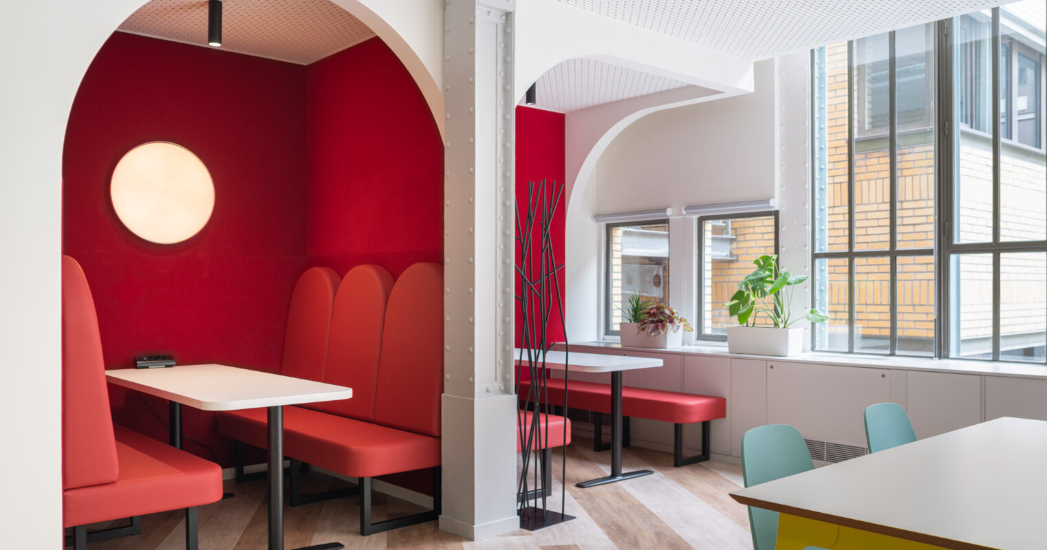 Modern communal space featuring bold red walls and curved archways that create intimate seating areas. Plush red booths provide comfort, while circular lighting adds warmth. Large windows allow natural light to flood the area, enhancing the vibrant color palette. Minimalist furnishings complement the contemporary aesthetic.