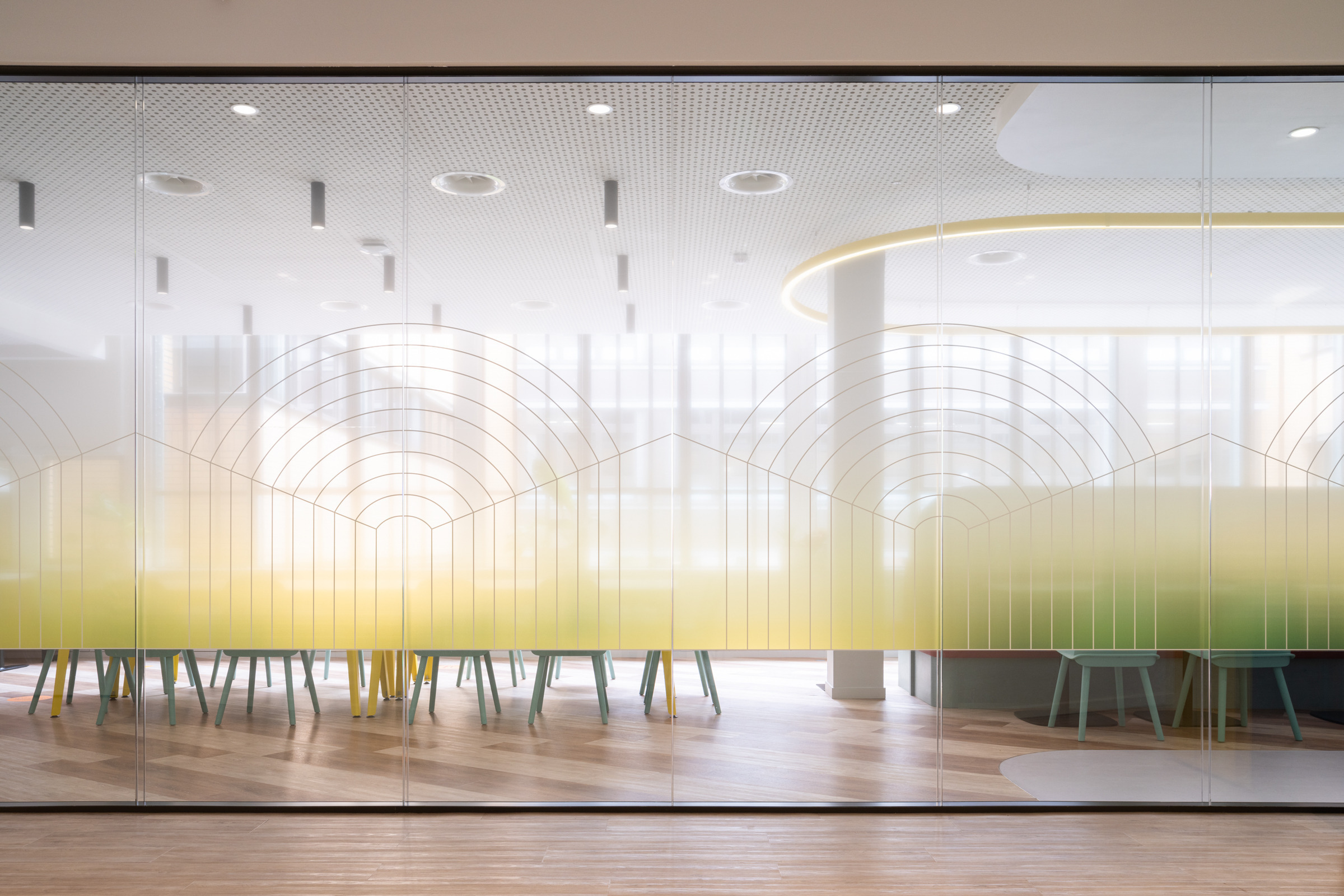 Frosted glass partitions showcase an innovative design featuring delicate curved patterns that transition from clear to a soft yellow hue. The spacious interior reveals colorful chairs and a modern table setup, creating a vibrant yet tranquil environment ideal for collaborative work and meetings. Natural light enhances the welcoming atmosphere.