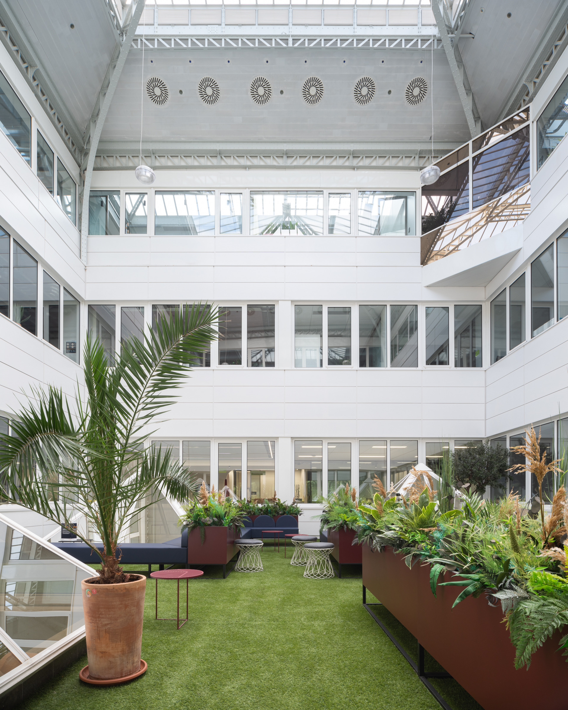 Bright, airy atrium featuring lush greenery and modern furnishings. Large glass windows allow natural light to flood the space, enhancing the open layout. Planters with tropical plants intermingle with sleek seating options. Architectural details include a high ceiling with unique lighting fixtures, creating an inviting atmosphere for collaboration.