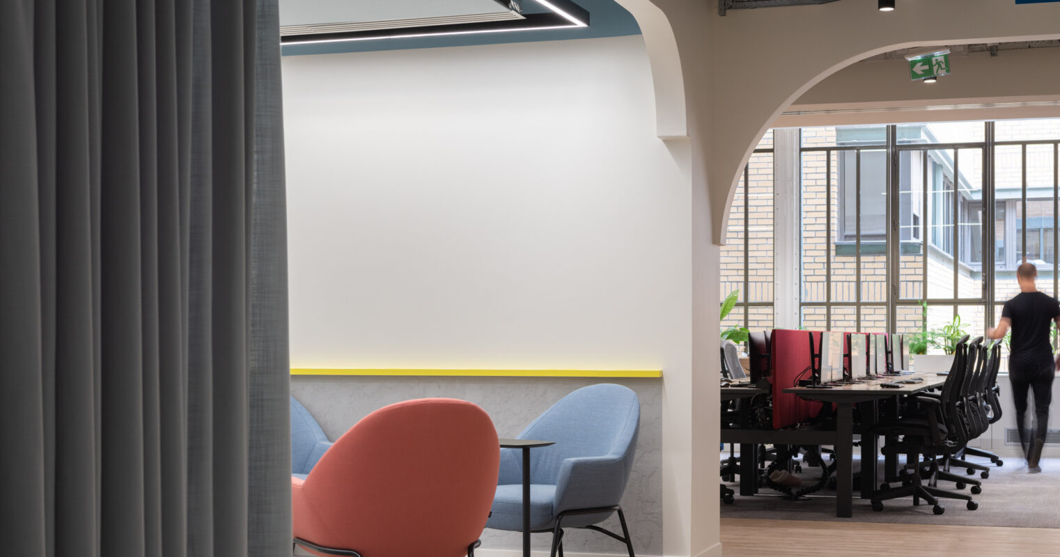 Modern office space featuring a blend of soft seating in coral and pastel blue, positioned near a workspace with sleek desks. Natural light filters through large windows, enhancing the airy atmosphere. Unique architectural arches create an inviting transition between collaborative and focused work areas, emphasizing functionality and comfort.