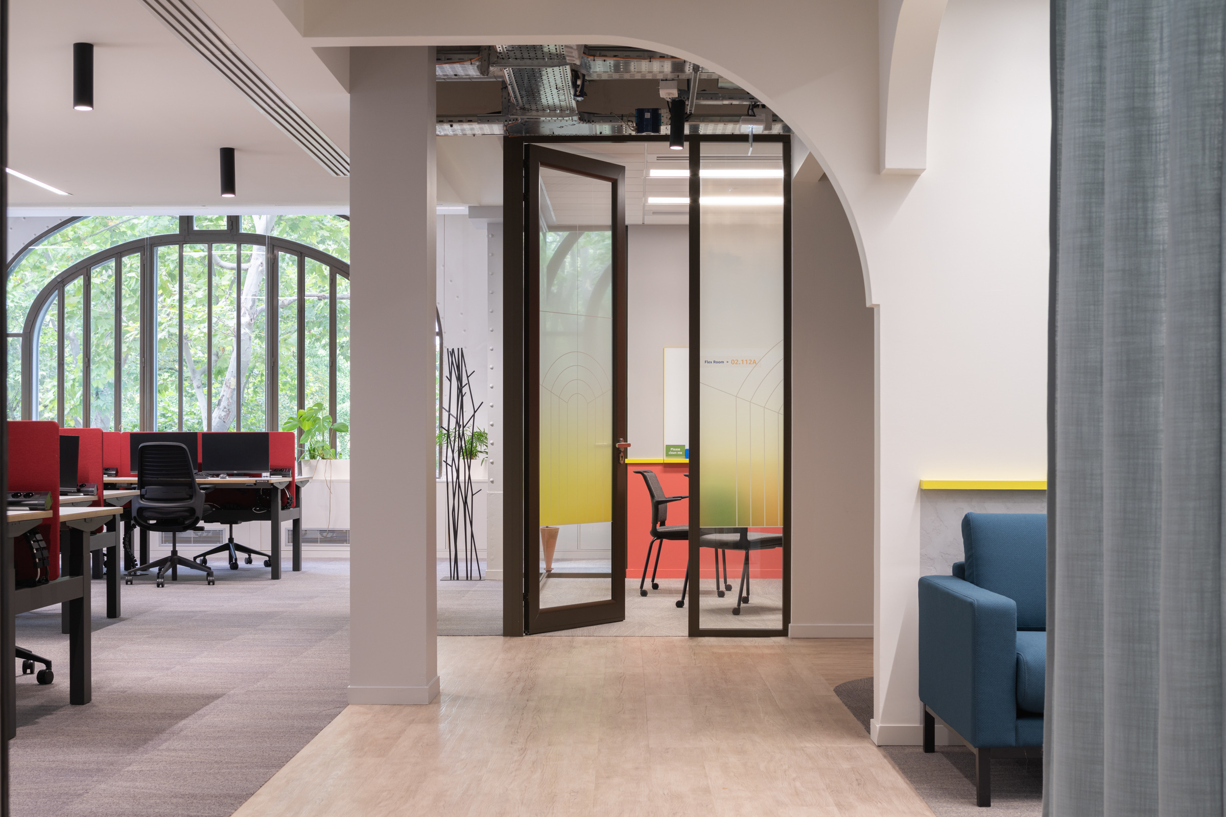 Modern office space featuring a blend of open and private areas. A glass-walled meeting room showcases gradient frosted glass, enhancing privacy while allowing natural light. The curved archway adds a soft architectural element, contrasting with sleek desks and ergonomic chairs in the background, promoting a functional yet inviting atmosphere.