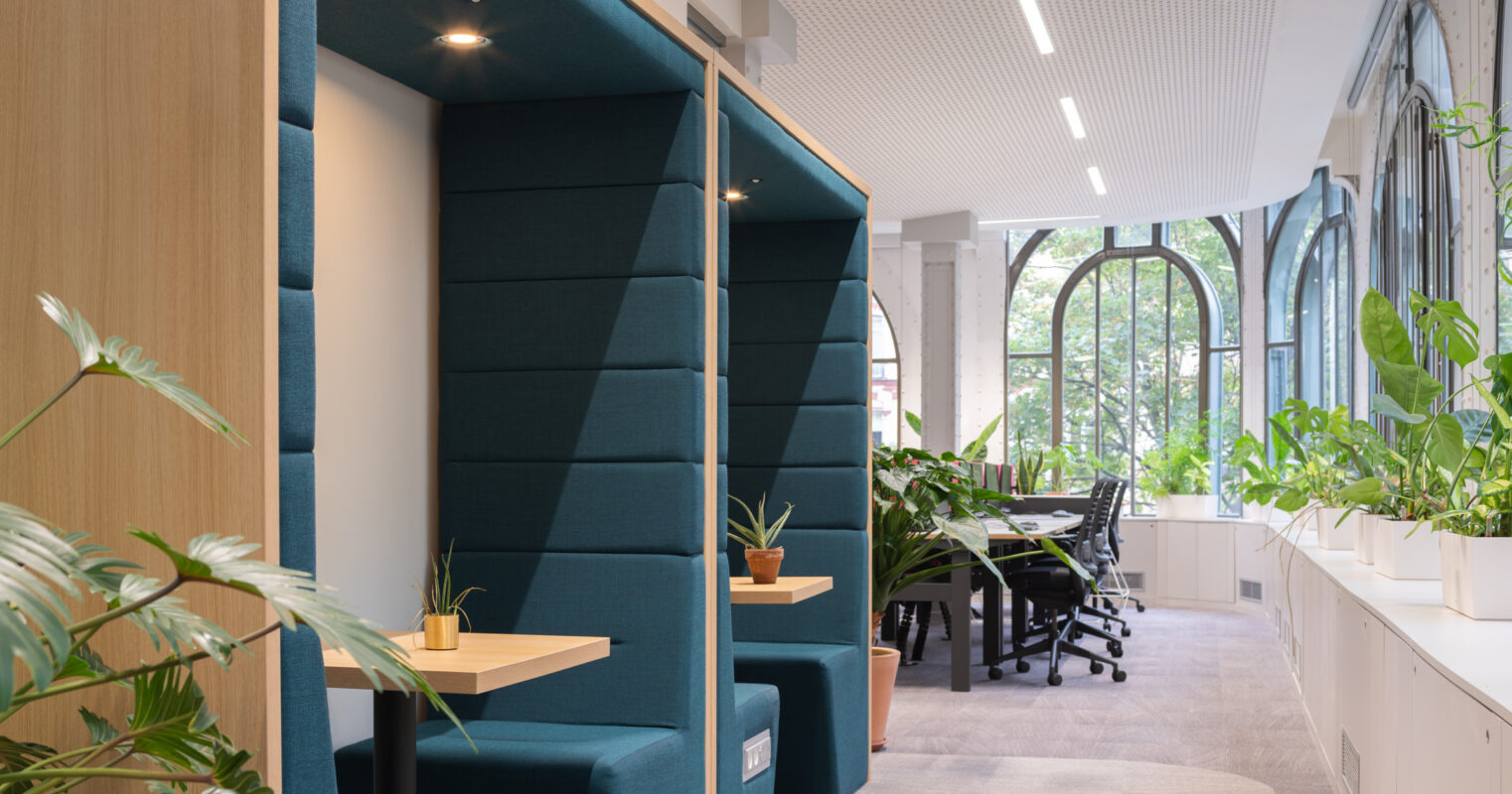 Modern office space featuring acoustic pods for privacy, adorned with deep teal upholstery. Lush greenery enhances the atmosphere, contrasting with the light-colored walls and carpet. Large windows invite natural light, fostering a productive environment. Ergonomic workstations are visible in the background, promoting collaboration and comfort.