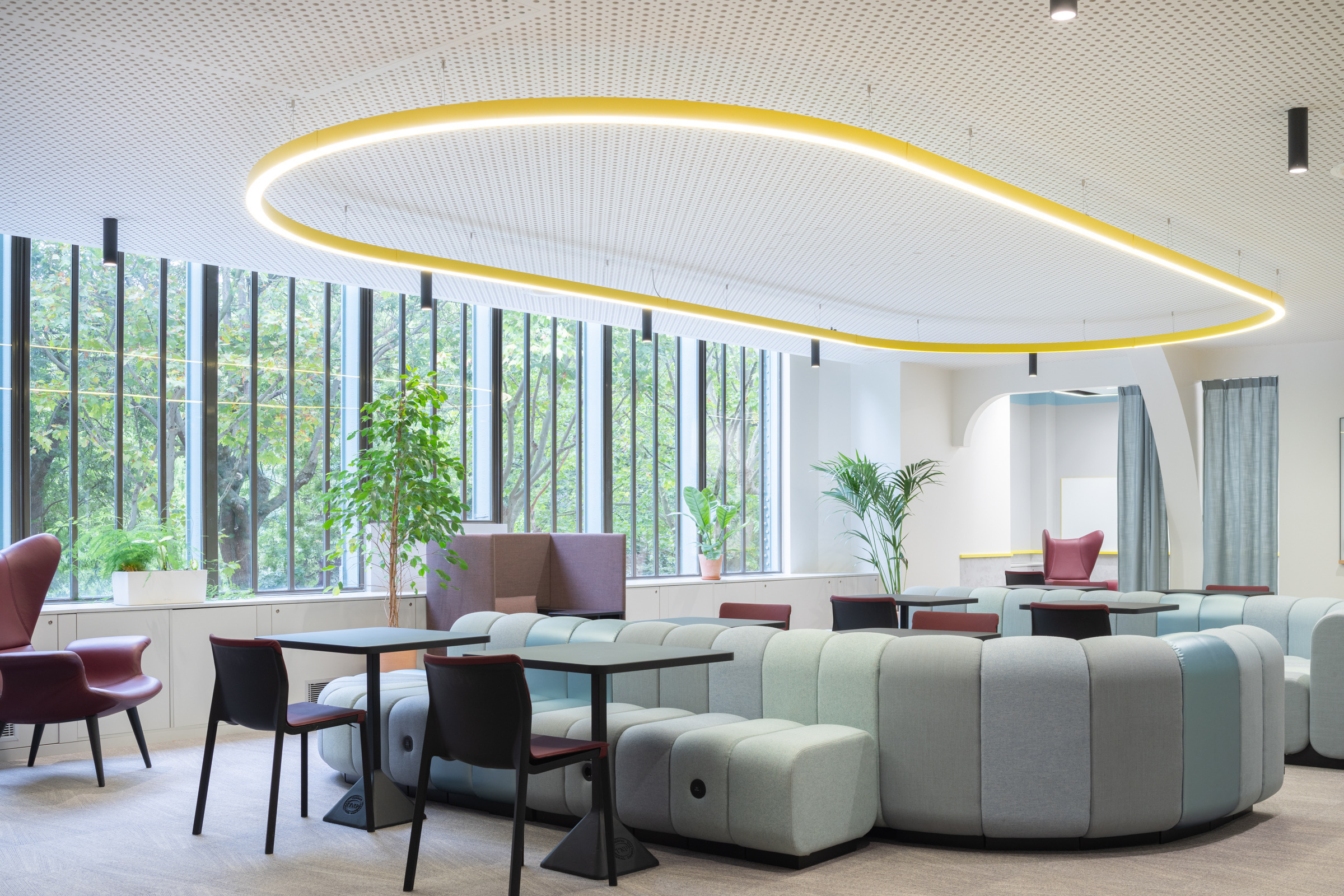 Contemporary coworking space featuring curved, modular seating in soft blue tones and rich burgundy accents. Large windows invite natural light, complemented by overhead lighting with a sleek, elongated design. Lush greenery adds vibrancy, creating a welcoming atmosphere that blends functionality with modern aesthetic appeal.