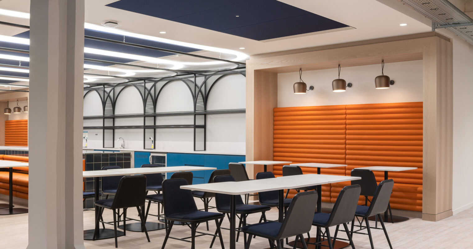 Modern dining area featuring sleek, black chairs and minimalist tables arranged around a vibrant orange acoustic wall panel. The ceiling showcases a unique grid of linear lighting, complemented by contrasting blue accents on cabinetry. Open, airy layout emphasizes contemporary design married with functionality, creating a welcoming atmosphere.