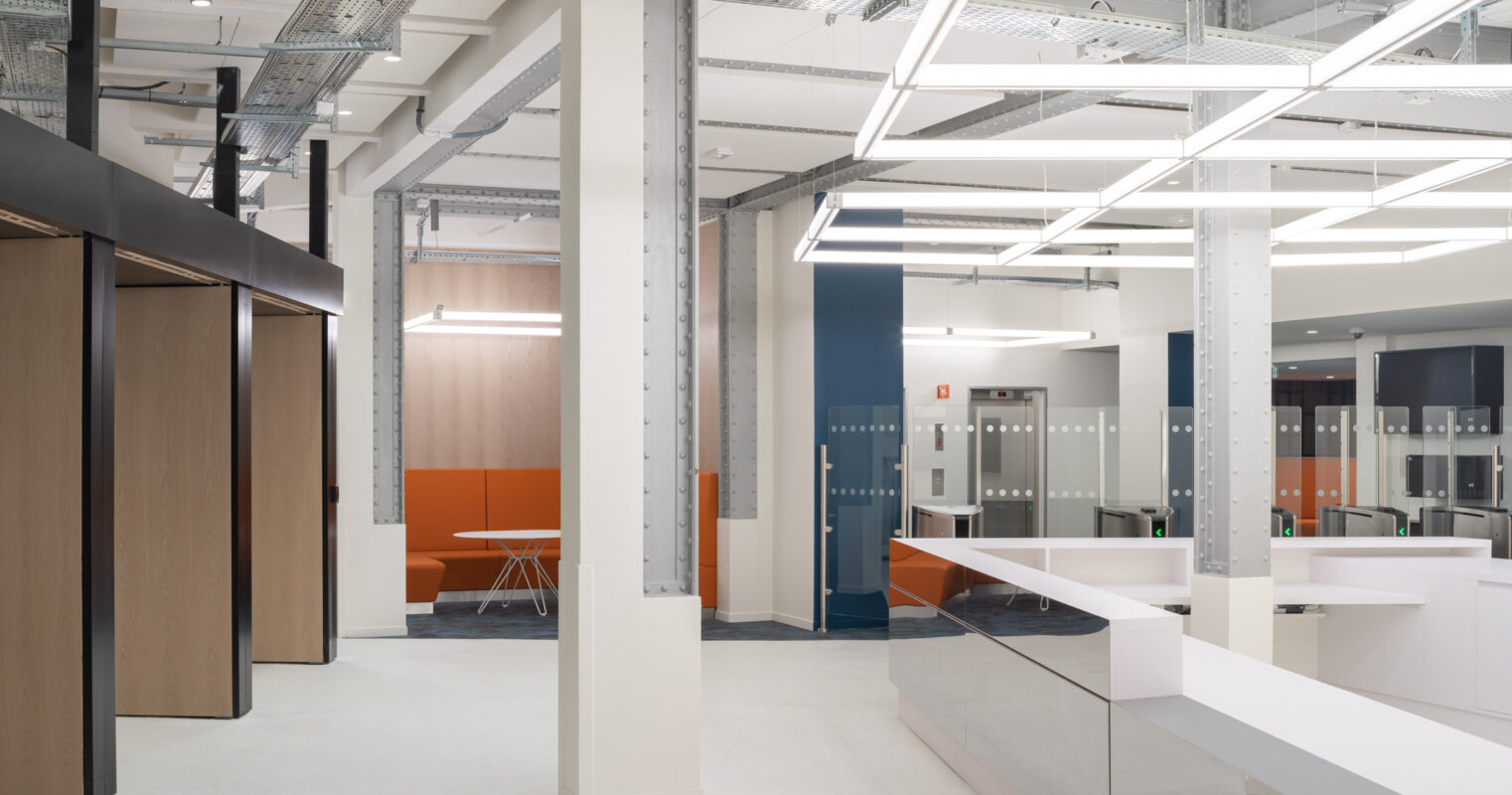 Modern office space featuring a blend of open collaboration areas and private meeting rooms. Sleek, minimalist furnishings complement exposed steel beams and industrial lighting. A spacious reception area showcases a contemporary front desk, with vibrant orange accents enhancing the overall professional atmosphere.