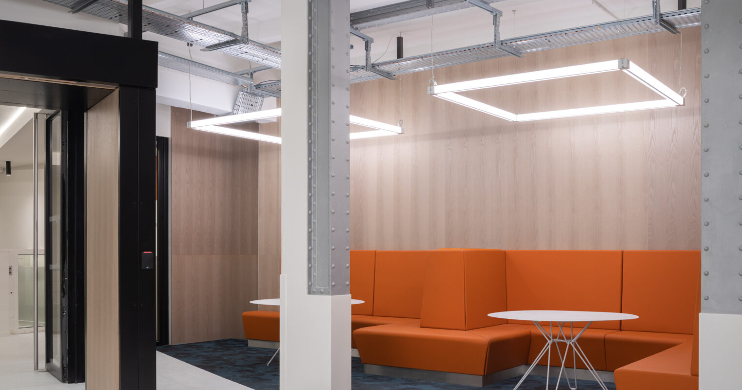 Modern collaborative workspace featuring warm orange seating and minimalist white tables. Exposed metal beams and linear light fixtures complement the wooden paneling, creating a balanced, inviting atmosphere. The design encourages both casual meetings and a relaxed working environment, showcasing contemporary interior design principles.