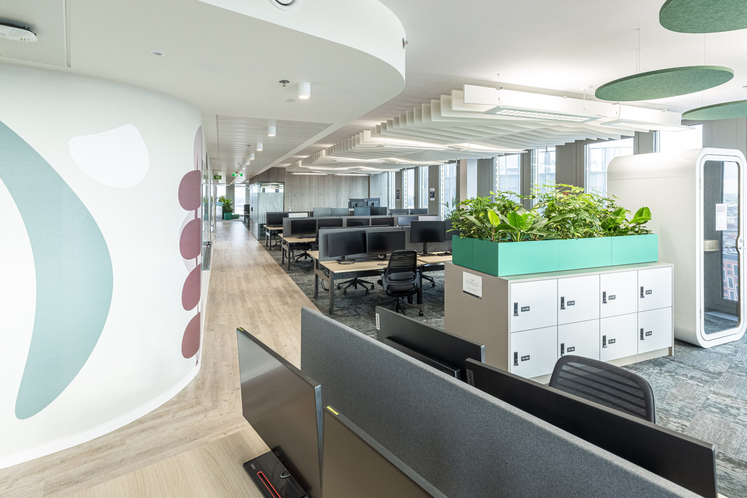 Modern office space featuring an open layout with sleek wooden flooring. The room is divided by a circular wall adorned with abstract patterns. Workstations include ergonomic chairs, dual monitors, and greenery-enhanced partitions. A soundproof pod for meetings is visible, promoting collaboration and privacy in the vibrant, well-lit environment.