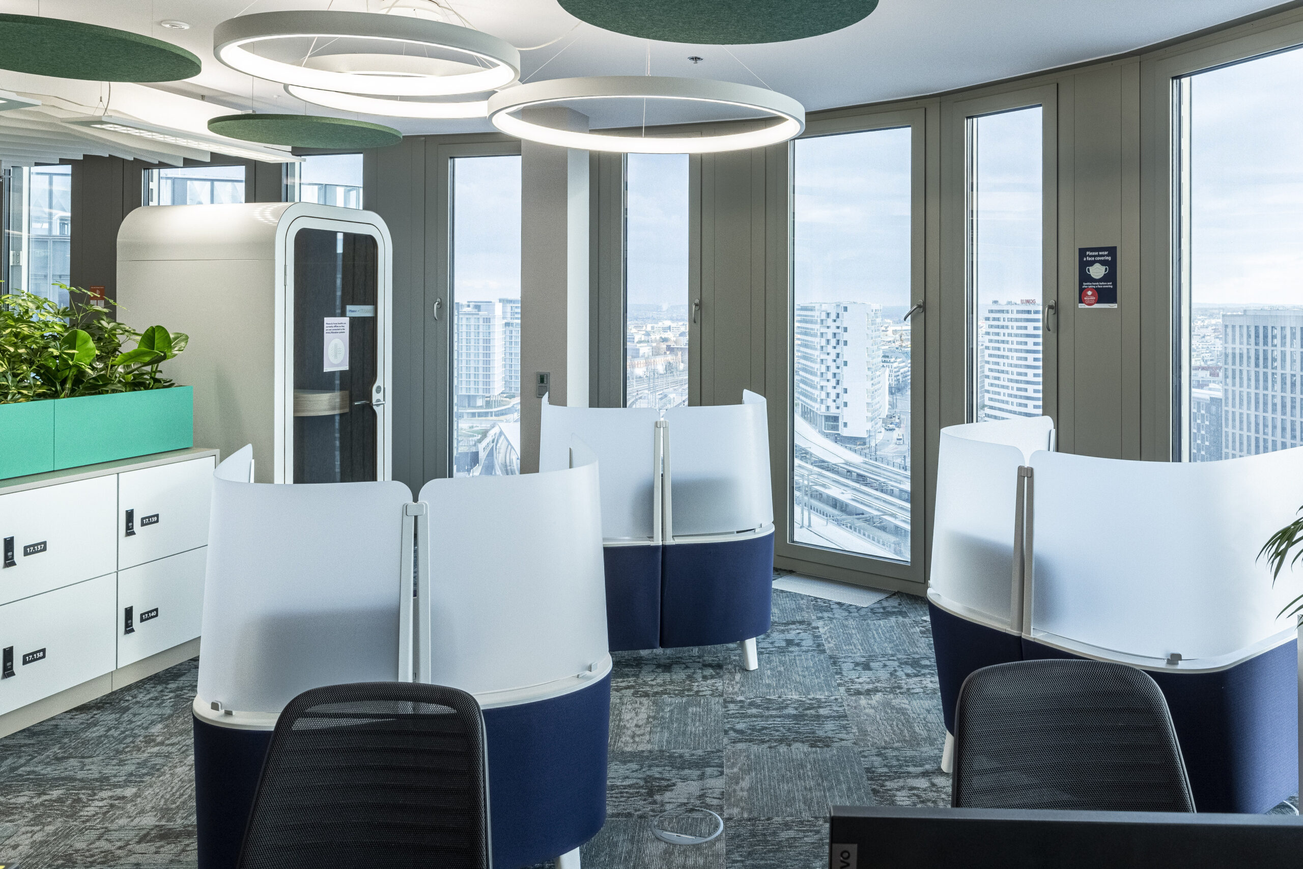 This modern office space features sleek, semi-private workstations with transparent backings, enhancing collaboration while maintaining privacy. Large windows provide abundant natural light and stunning city views. Greenery along the walls adds a touch of nature, promoting a healthy work environment. Innovative lighting fixtures create an inviting atmosphere.