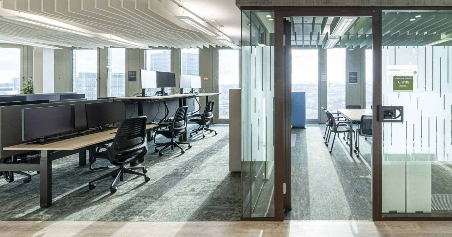 Natural light floods a contemporary office space featuring sleek workstations with ergonomic chairs positioned along a minimally designed open floor plan. Glass partitions create private areas while maintaining an airy feel. The textured flooring complements the modern aesthetic, enhancing both functionality and visual interest in the workspace.