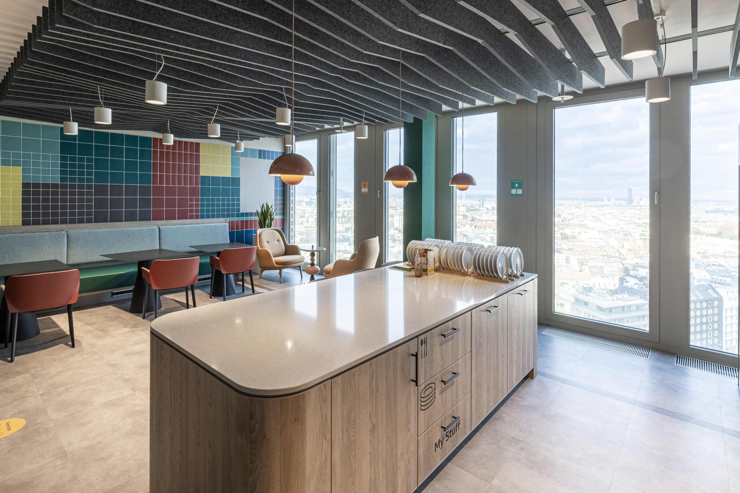 Bright and airy modern kitchen featuring sleek cabinetry and a large island with a polished countertop. Stylish pendant lights hang from a textured ceiling, while a colorful tiled wall adds vibrancy. A dining area with plush seating offers city views, enhancing the contemporary, inviting atmosphere.