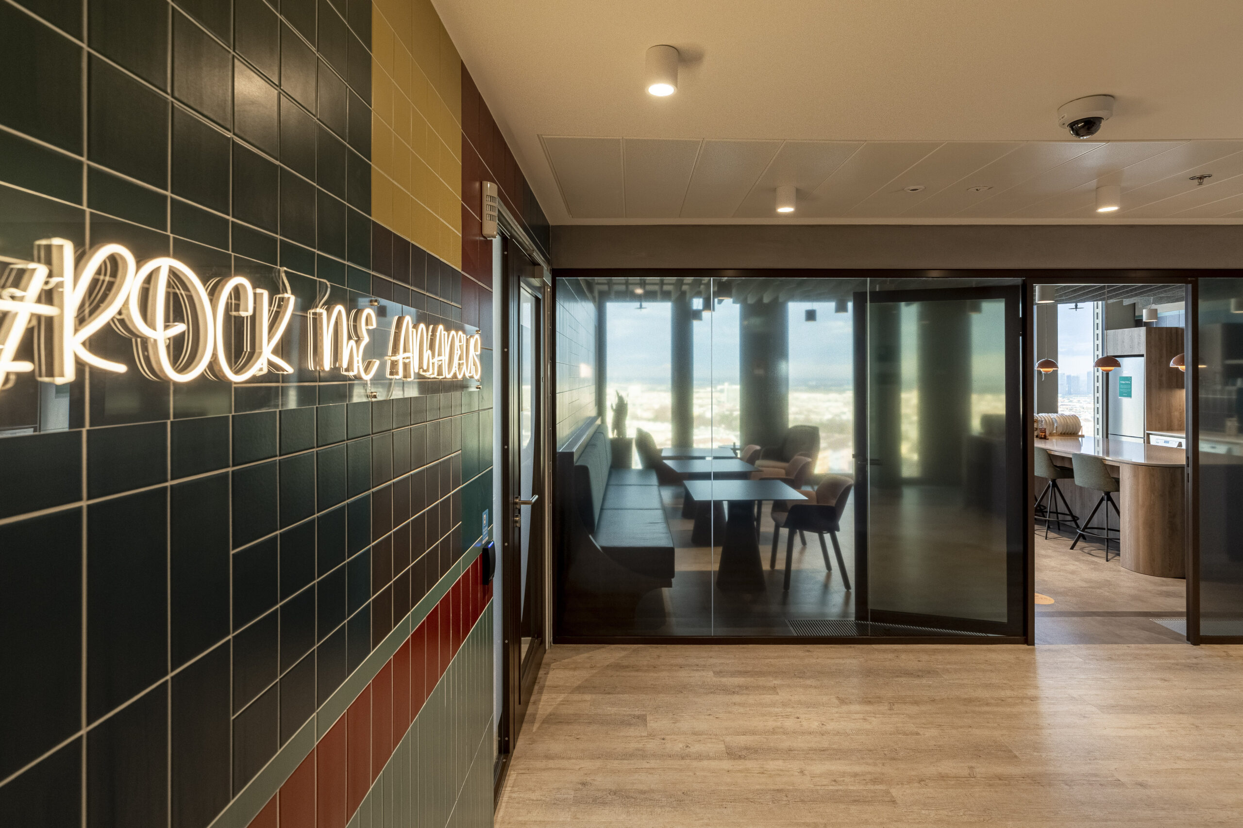 A vibrant interior corridor features contemporary wall tiles in various colors, creating visual interest. A glowing neon sign reading "#ROCK ME HAPPY" adds a playful touch. Beyond the glass doors, a stylish conference room showcases modern furniture and panoramic views of the skyline, enhancing the workspace atmosphere.