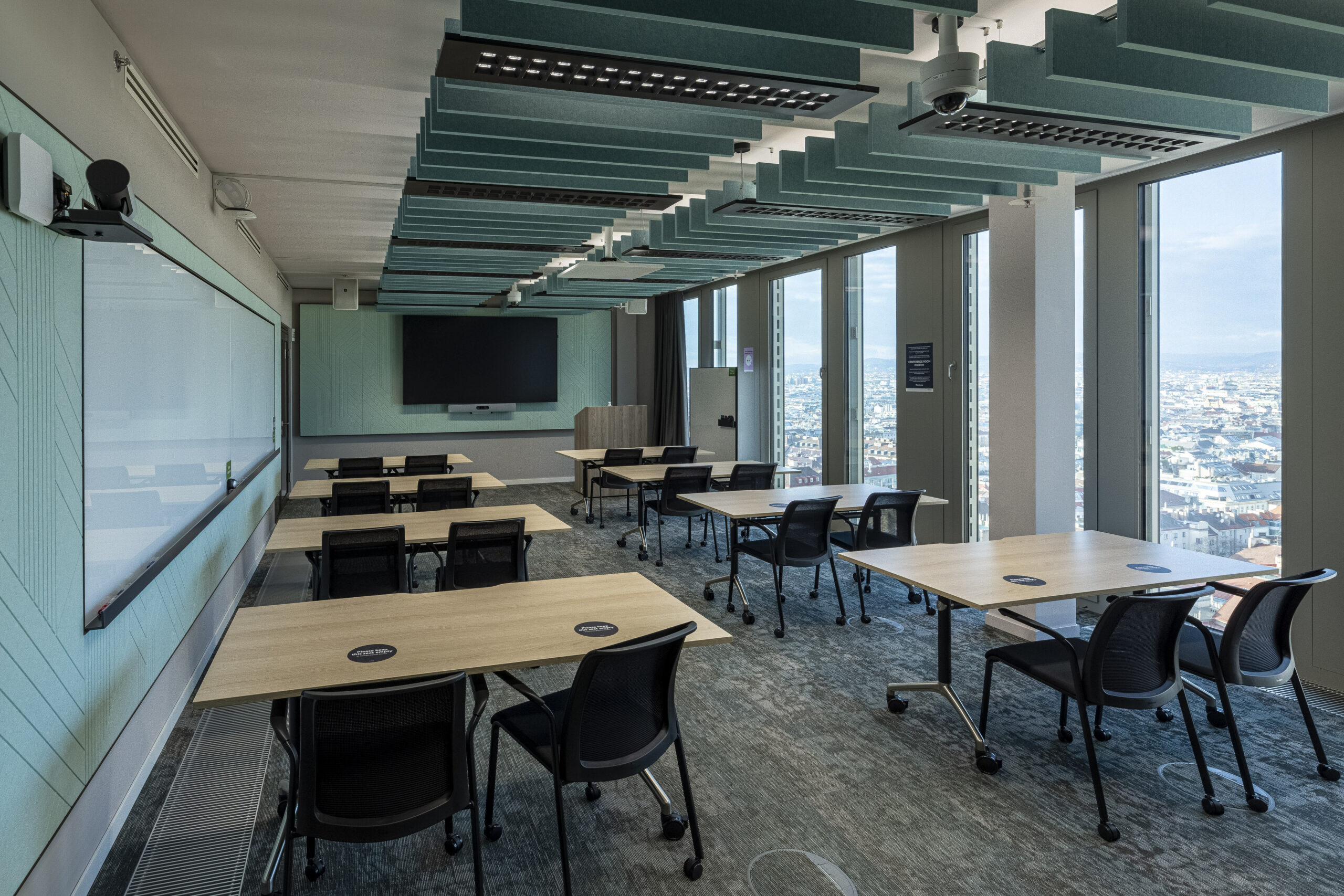 A modern conference room featuring modular seating and flexible workspace arrangements. Light-colored wooden tables with integrated power outlets complement the acoustic ceiling panels, enhancing sound quality. Large windows offer expansive city views, while the light teal walls create a fresh, professional atmosphere conducive to collaboration and creativity.