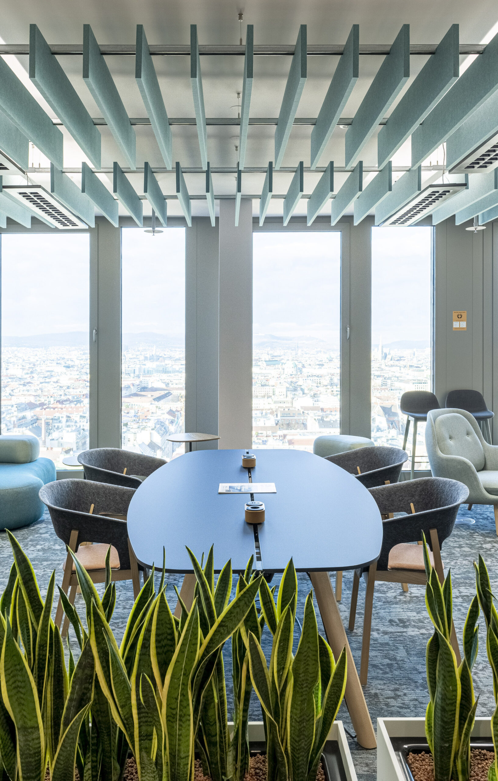 Modern office space featuring a sleek blue conference table surrounded by ergonomic chairs. Above, innovative ceiling beams provide structural interest while filtering natural light. Lush green plants in the foreground enhance the workspace's ambiance, promoting a sense of well-being and connection to nature amid a panoramic city view.