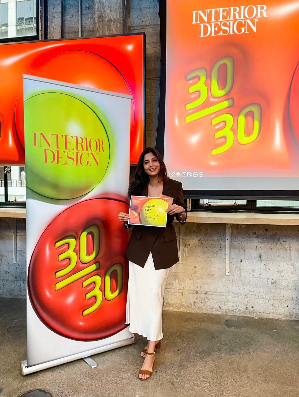 A young woman stands confidently in front of a modern presentation backdrop showcasing the "Interior Design 30/30" event. She holds a colorful booklet, dressed in a stylish brown blazer and white skirt. The venue features sleek industrial elements, combining contemporary aesthetics with a professional ambiance, enhancing the event's focus on innovative design.