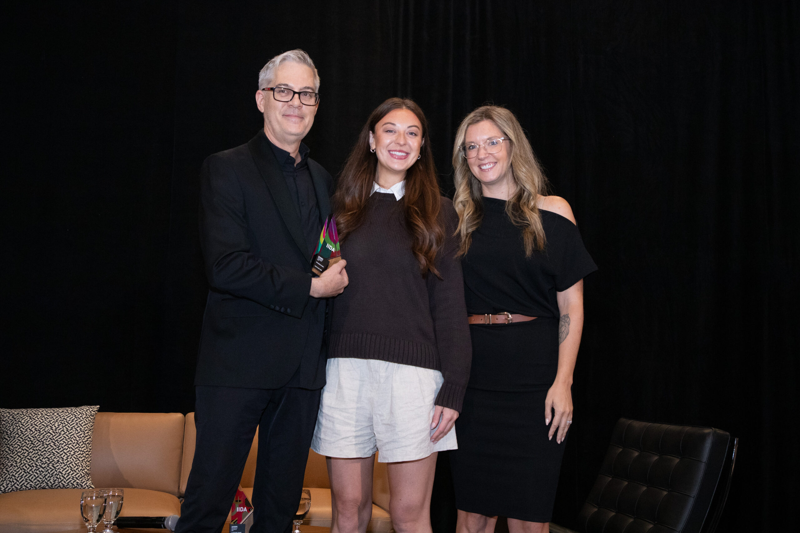 Three professionals stand together in a studio setting, smiling warmly. The first, wearing glasses and a black suit, exudes authority. The second, dressed casually in a dark sweater and shorts, showcases a contemporary, youthful aesthetic. The third, in a chic black dress, radiates sophistication, contributing to a collaborative atmosphere.