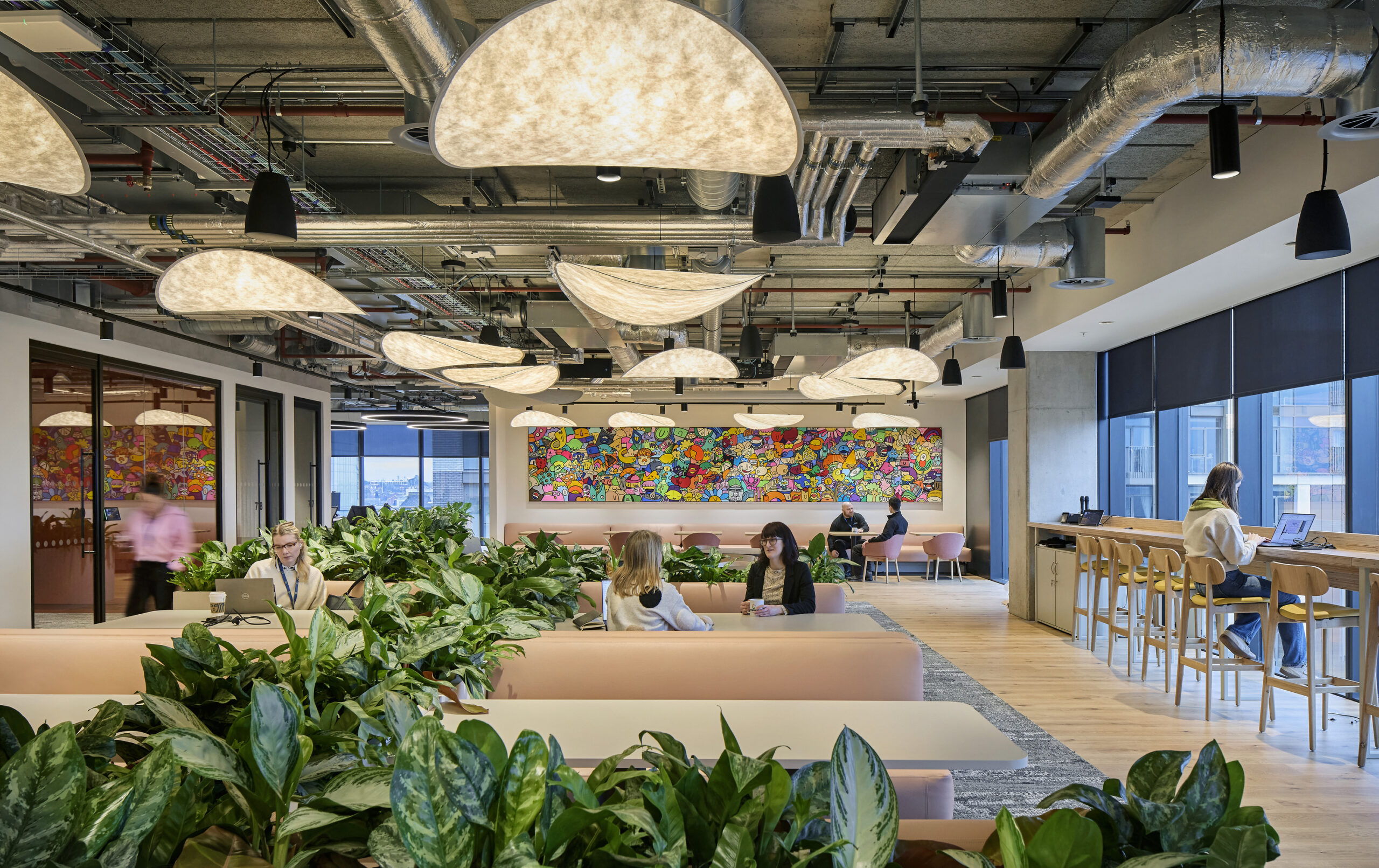 Open office space featuring modern, curved pendant lighting that casts a warm glow. Abundant indoor plants create a lively atmosphere, while a vibrant mural adds a pop of color. Comfortable seating areas encourage collaboration, complemented by sleek, minimalist furniture and large windows allowing natural light to enhance productivity.