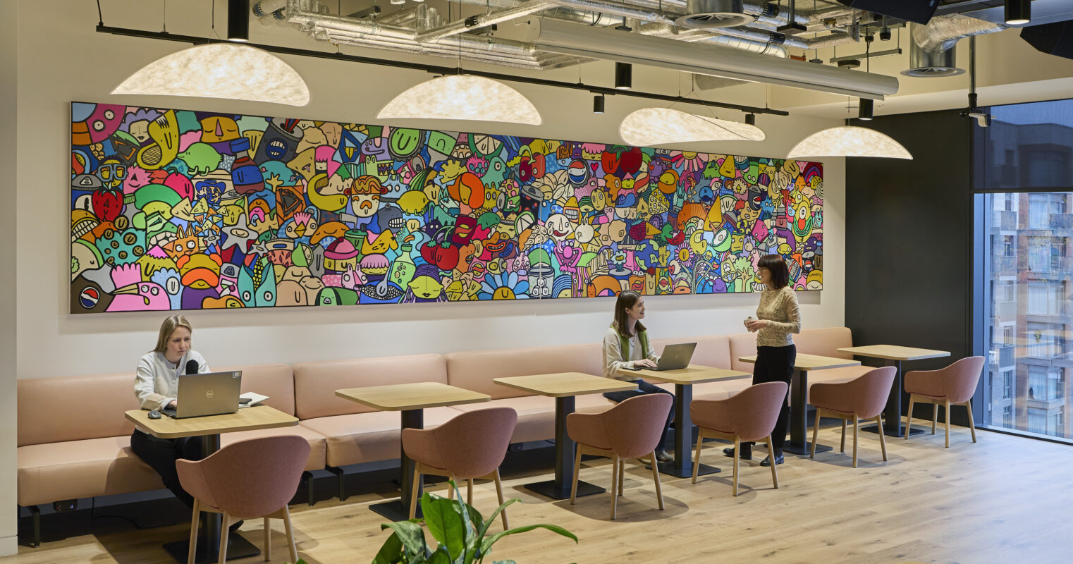 A vibrant café space features a whimsical mural, filled with cartoonish characters and colorful designs, creating an energizing focal point. Soft pink seating aligns with minimalist wooden tables, while stylish pendant lights illuminate the area. Lush greenery complements the modern aesthetic, inviting collaboration and relaxation.