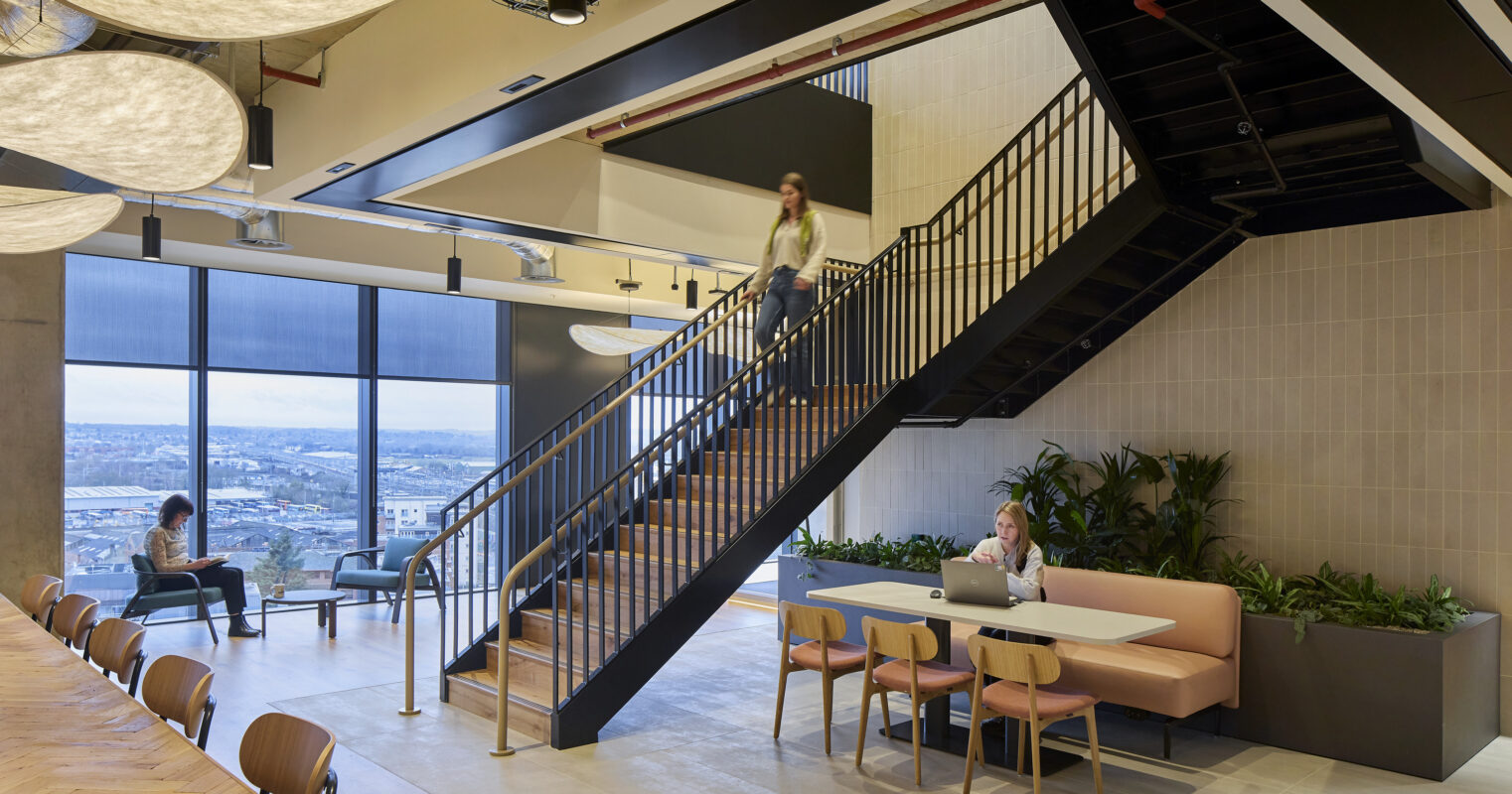 This modern interior features a multi-level workspace highlighting a striking staircase with a wooden handrail, leading to a bright area adorned with large windows. Soft, organic pendant lights hang overhead, while comfortable seating and greenery provide a refreshing balance, fostering a collaborative and inviting environment.