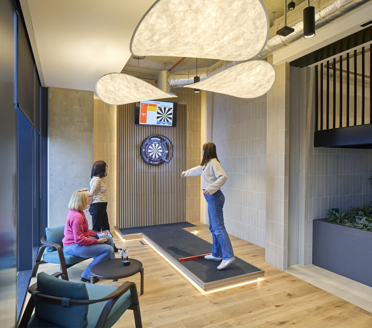 A modern recreational space featuring a dartboard on a textured wooden accent wall. Soft, cloud-like pendant lights illuminate the area, enhancing its inviting atmosphere. Two seated women observe, while a third, poised to throw, stands on a sleek platform. Natural wooden flooring complements the contemporary design elements.