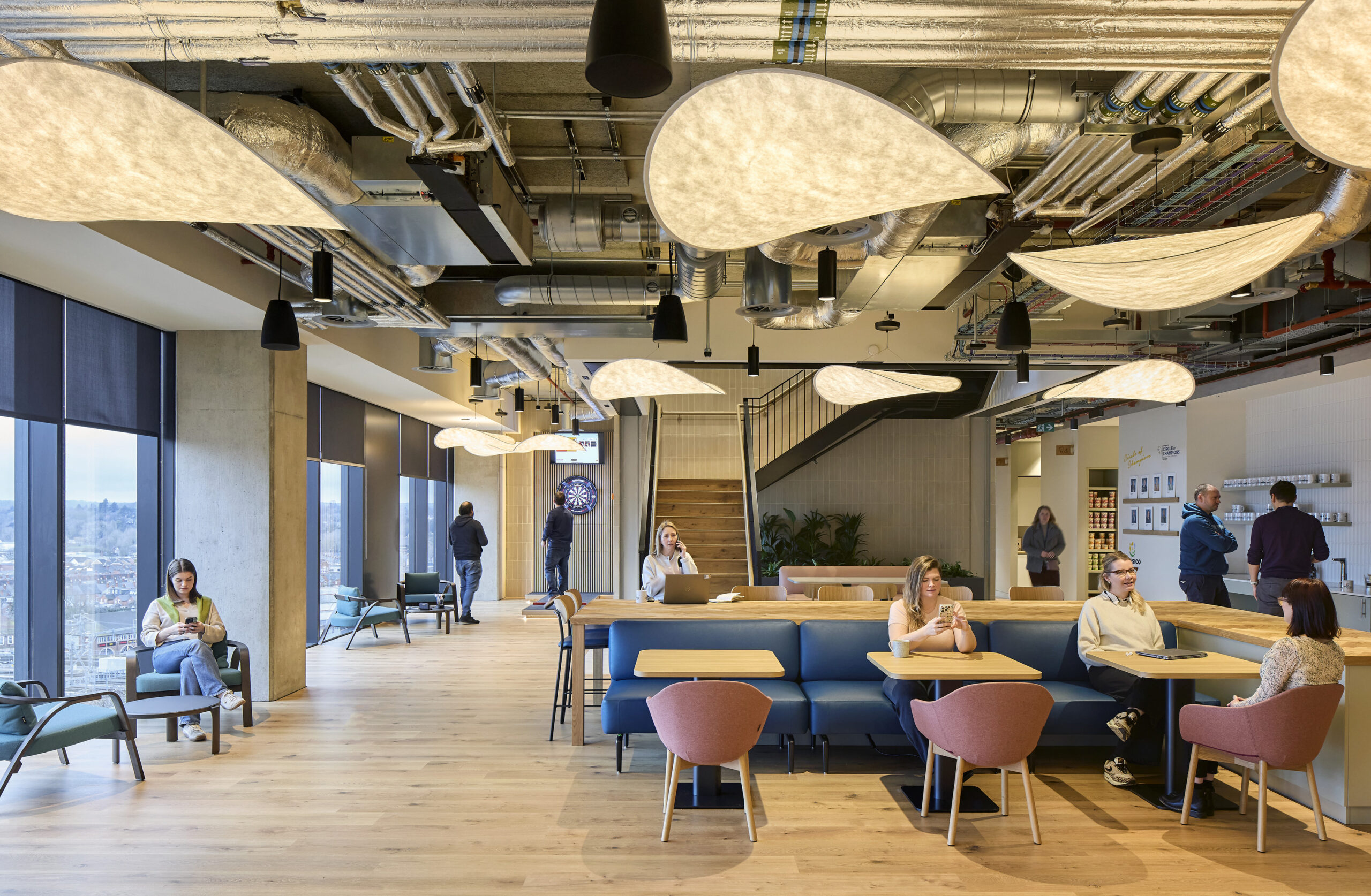 Modern office interior featuring a blend of collaborative and relaxed spaces. Unique, organic-shaped light fixtures complement exposed ductwork, highlighting an industrial aesthetic. Natural wood flooring and soft seating in muted colors create a welcoming atmosphere for employees. Large windows offer expansive views, enhancing the space's openness.