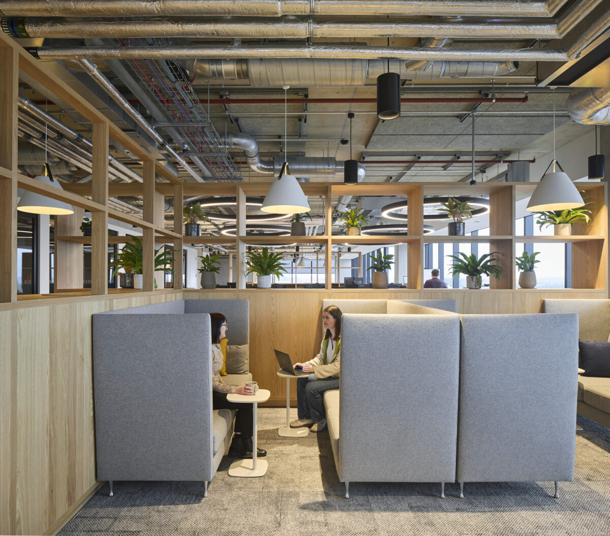 Contemporary office space featuring modular seating with high backrests, promoting privacy. Warm wooden panels and lush greenery enhance the inviting atmosphere. Industrial-style elements, including exposed ductwork and pendant lighting, contrast with the soft furnishings. Workstations are designed for collaboration and comfort, fostering productivity and creativity.