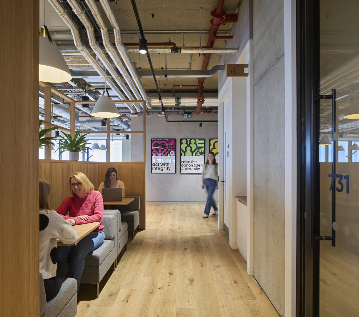 Modern office space features collaborative seating arrangements with cozy gray upholstery, wooden accents, and ambient lighting. A well-designed layout encourages interaction, highlighted by vibrant wall art conveying motivational messages. Exposed ductwork and large windows contribute to an open, contemporary atmosphere, enhancing productivity and creativity among occupants.