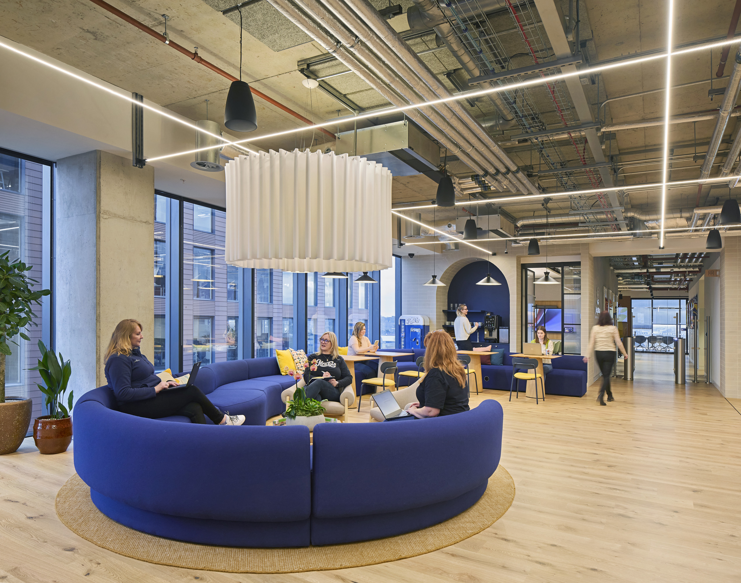 Spacious modern office featuring a circular blue sofa with bright cushions, encouraging collaboration. Natural light floods the room through large windows, highlighting industrial design elements such as exposed ceilings and pendant lighting. A relaxed atmosphere is enhanced by plants and varied seating arrangements for diverse work styles.