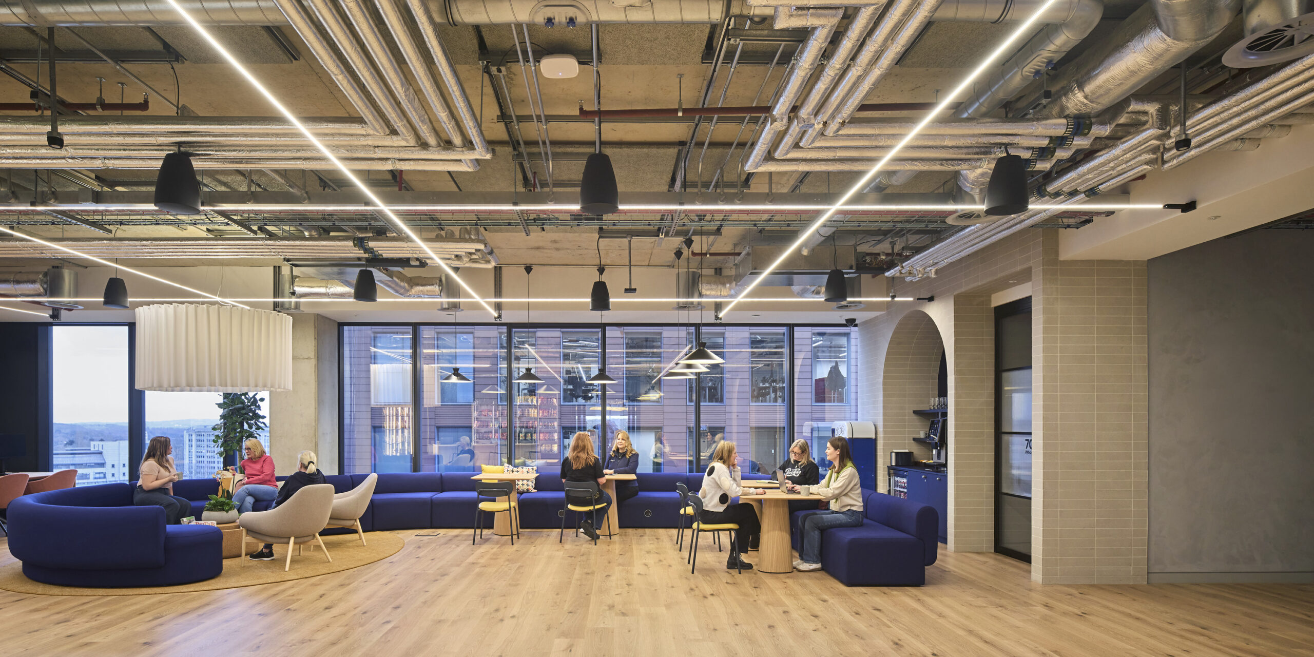 A contemporary office design features an open layout with a striking mix of casual seating areas and collaborative workspaces. Curved blue couches and light wood tables create an inviting atmosphere. Industrial elements like exposed ductwork and sleek light fixtures enhance a modern aesthetic, promoting both functionality and comfort.