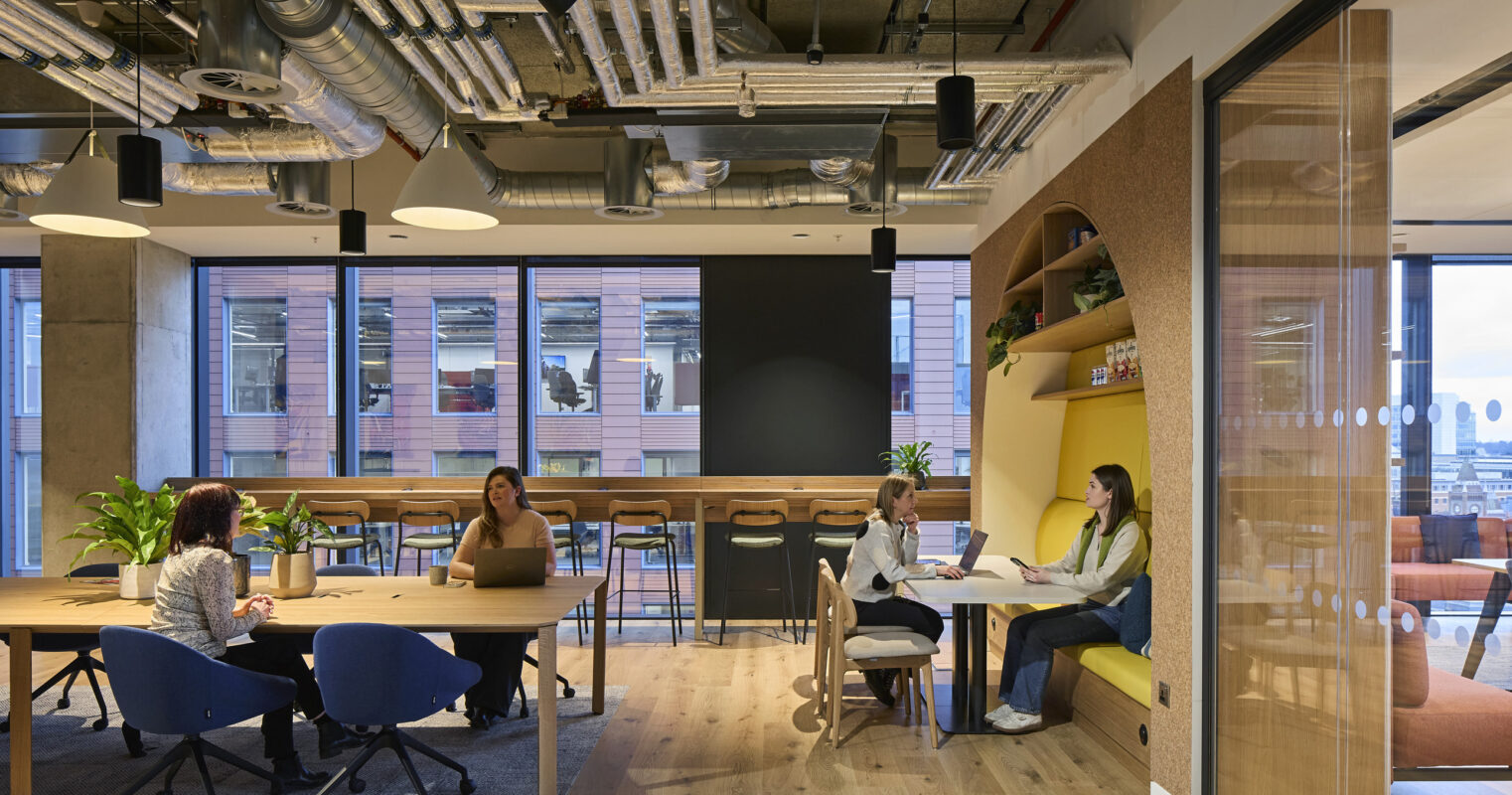 An open-plan office space features a blend of collaborative and casual seating areas. Modern wooden furniture complements the warm tones of natural light from large windows, while decorative plants enhance the inviting atmosphere. Distinctive arch-shaped shelving adds visual interest, emphasizing functional design.
