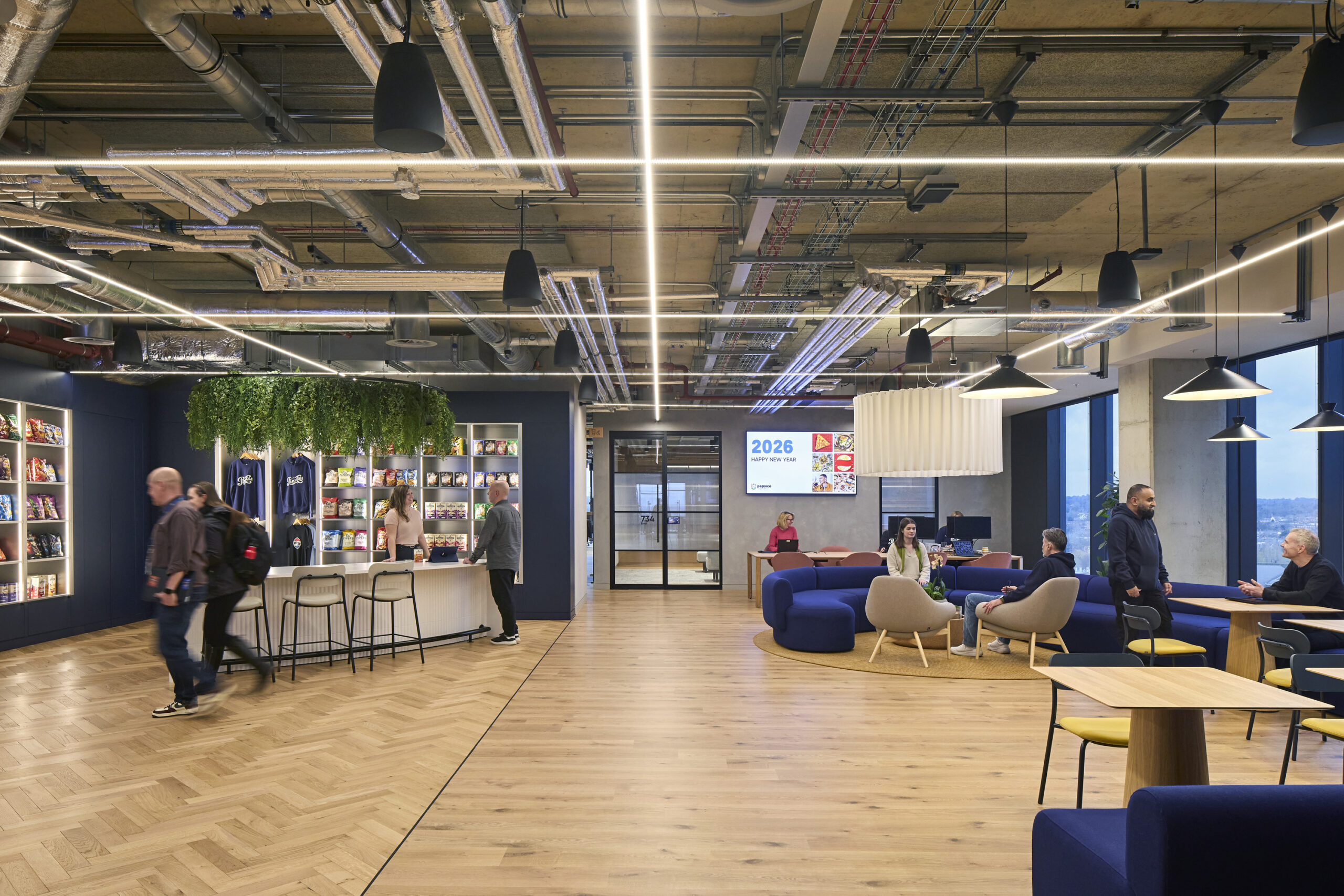 A modern office space featuring a vibrant, inviting layout. The wooden herringbone flooring contrasts with industrial ceiling beams and exposed ducts. A green plant installation cascades over a snack bar, while contemporary furniture promotes collaboration in the lounge area, illuminated by stylish overhead lighting and large windows.