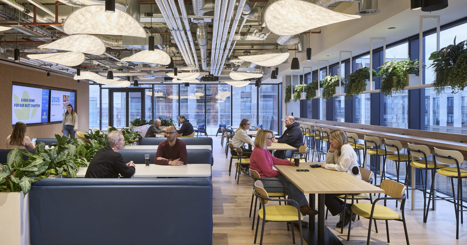 A contemporary office space features modern furnishings with comfortable blue seating arrangements and wooden tables, promoting collaboration. Unique, large light fixtures imitate organic shapes, while abundant greenery enhances the atmosphere. Floor-to-ceiling windows maximize natural light, offering a vibrant environment conducive to productivity and social interaction.