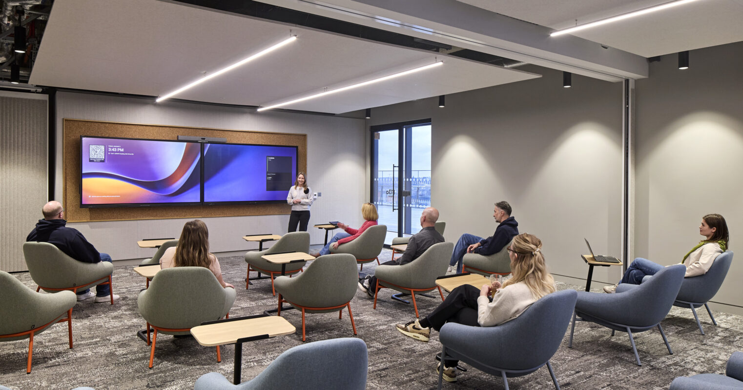 A modern collaborative workspace features a sleek, dual-screen display mounted on a textured wall, enhancing engagement during presentations. Curved, upholstered chairs with orange accents surround minimalist wooden tables, promoting comfort and interaction. Large windows provide ample natural light, creating an inviting atmosphere for meetings and discussions.