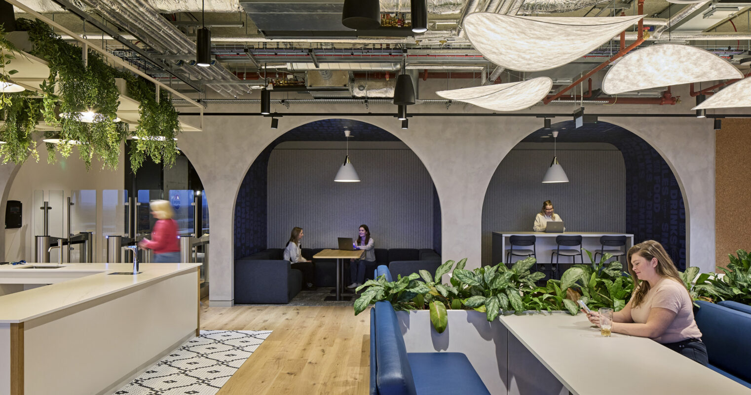 Innovative office collaboration space featuring large archways, soft seating, and abundant greenery. Natural light filters through the ceiling, complemented by modern pendant lighting. Design elements include textured walls and a blend of open and intimate areas, fostering creativity and interaction among team members throughout the environment.