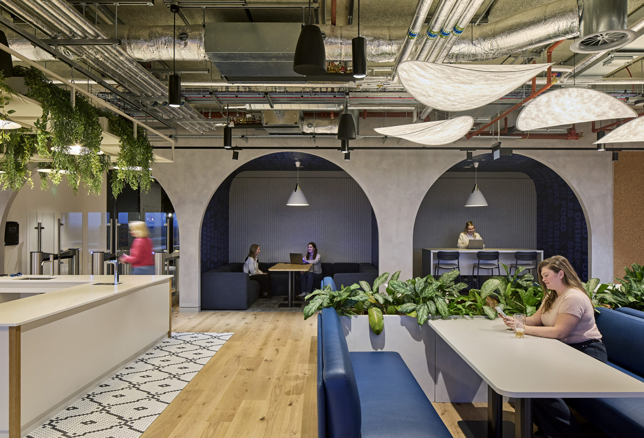 Innovative office collaboration space featuring large archways, soft seating, and abundant greenery. Natural light filters through the ceiling, complemented by modern pendant lighting. Design elements include textured walls and a blend of open and intimate areas, fostering creativity and interaction among team members throughout the environment.