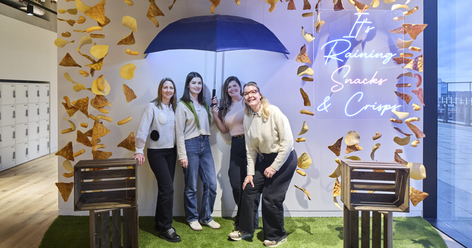 Vibrant interior space featuring a playful snack-themed design. Women pose under a bold blue umbrella surrounded by suspended snack decorations and a neon sign reading "It's Raining Snacks & Crisps." Two rustic wooden crates and realistic grass carpet enhance the whimsical atmosphere, promoting a fun, casual environment.