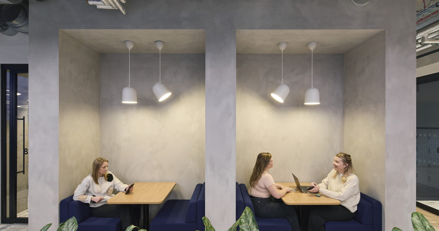 Two individuals engage in conversation within a cozy, modern nook featuring soft blue upholstered benches and warm wooden tables. Soft, rounded pendant lights illuminate the space, surrounded by subtle greenery. The textured grey walls contribute to a stylish yet functional workspace atmosphere, encouraging collaboration and relaxation.