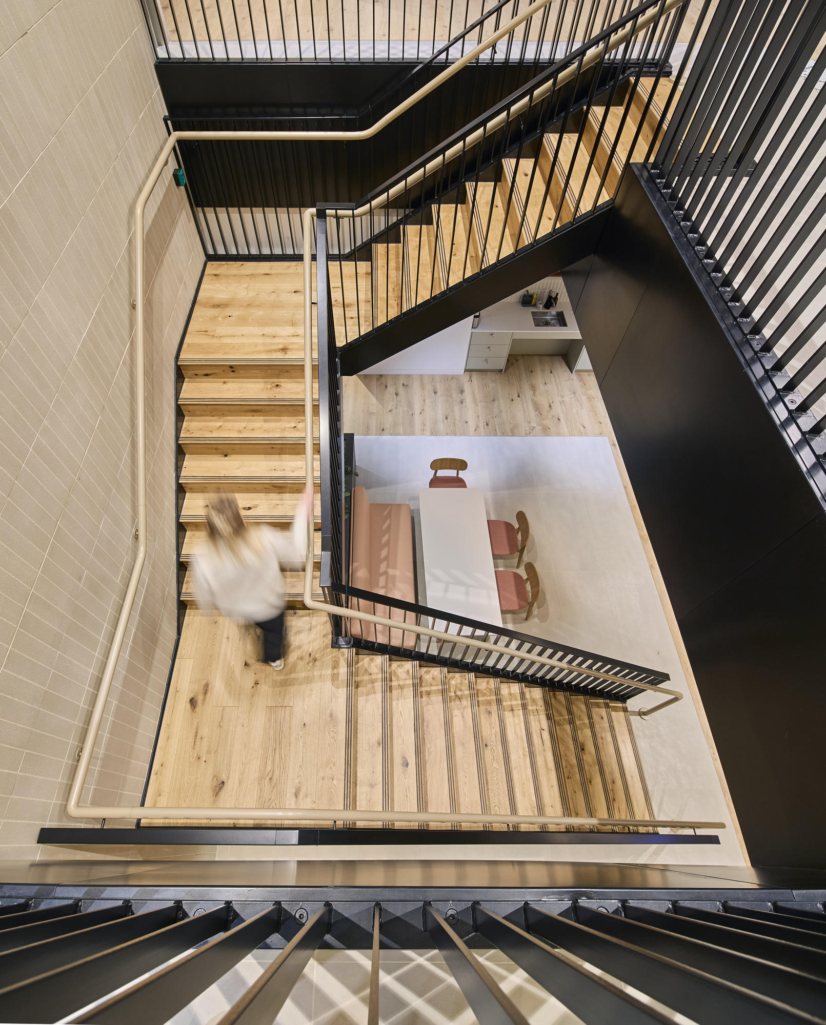 A modern staircase featuring wooden treads and sleek black railings, leading to an open area below. The space is defined by neutral tones, natural light, and minimalist furnishings, including a pink sofa and a white table, promoting a contemporary and inviting atmosphere.