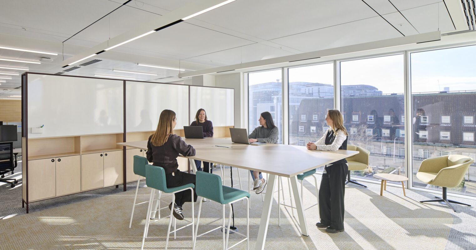 A bright, modern conference room features a large, minimalist wooden table surrounded by colorful bar stools. Five professionals engage in discussion while utilizing laptops. Large windows offer views of the cityscape, and natural light enhances the contemporary design, promoting collaboration and creativity in the workspace.
