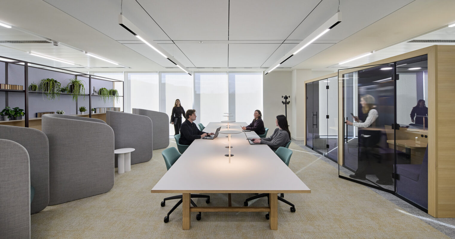 A stylish, modern office space featuring a long white table surrounded by ergonomic chairs. Textured gray acoustic panels create private workstations, while green plants add a touch of nature. Large windows provide ample natural light, enhancing productivity and well-being in the work environment.
