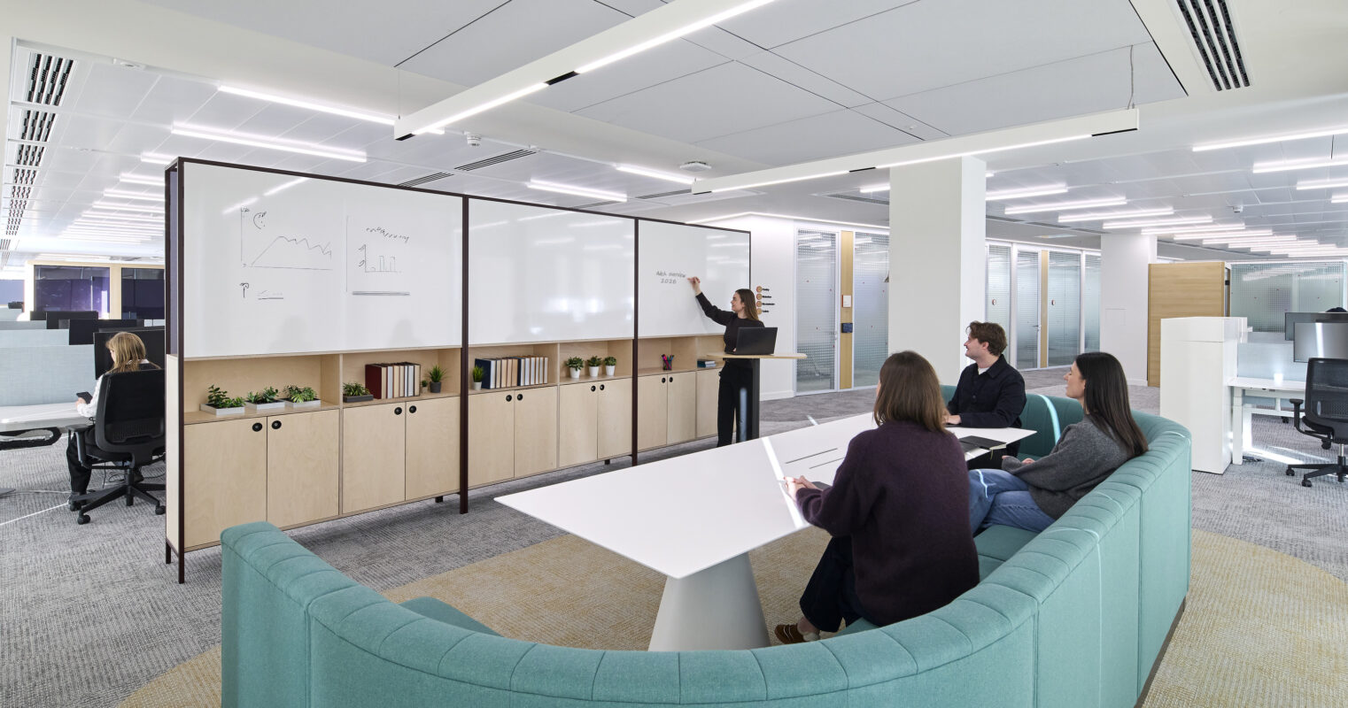 A modern office space featuring a collaborative area with a curved teal sofa. A presenter engages colleagues using a whiteboard, while those seated write notes at a sleek white table. The design emphasizes open communication, natural light, and multifunctional furniture, creating an inviting and productive atmosphere.