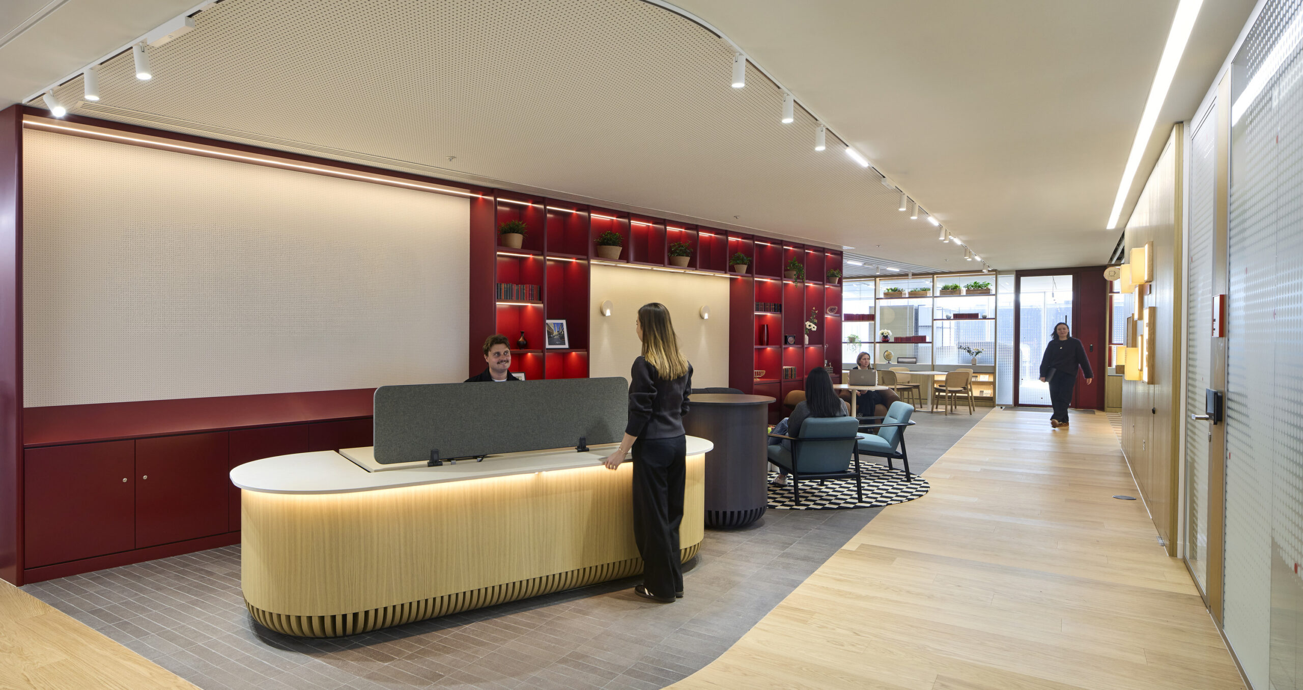 Reception area features a sleek, rounded desk with integrated lighting, set against a backdrop of vibrant red shelving accented by greenery. The open layout promotes collaboration, showcasing modern furnishings such as comfortable seating and a striking black and white patterned rug, enhancing both functionality and aesthetic appeal.