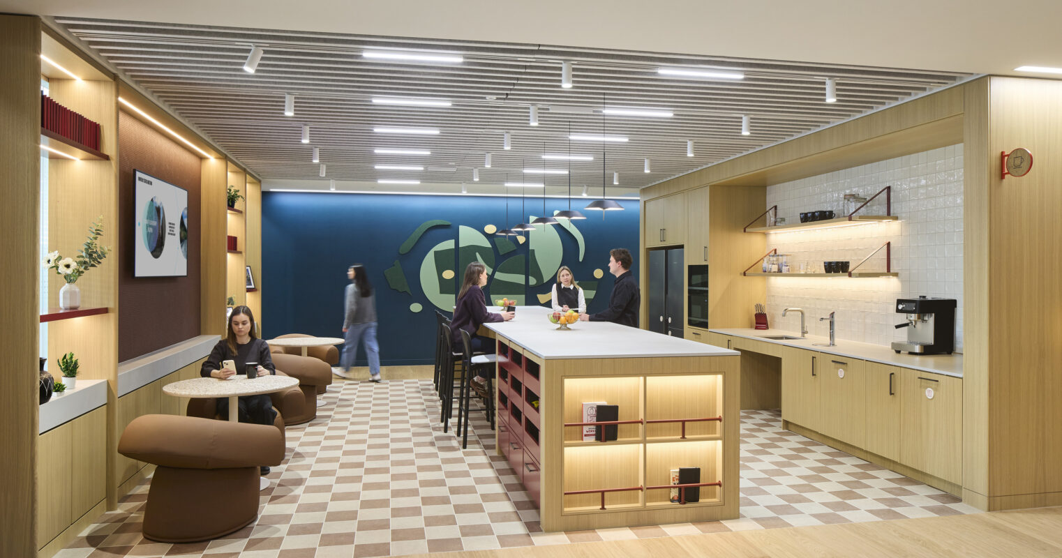 Bright, open workspace featuring a communal kitchen area with contemporary wood cabinetry and sleek appliances. Comfortable seating arrangements in warm tones create informal gathering spots. A vibrant blue accent wall adorned with abstract shapes adds visual interest, while a checkered floor design brings a playful element to the modern aesthetic.